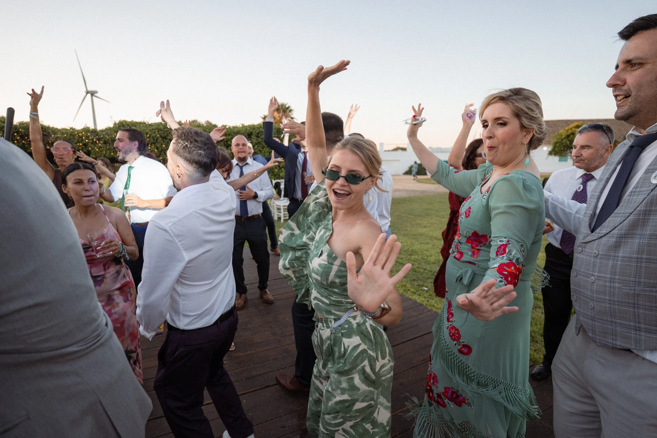 Grupo de personas bailando y disfrutando en una fiesta al aire libre con vista al mar y un aerogenerador en el fondo, durante el atardecer.
