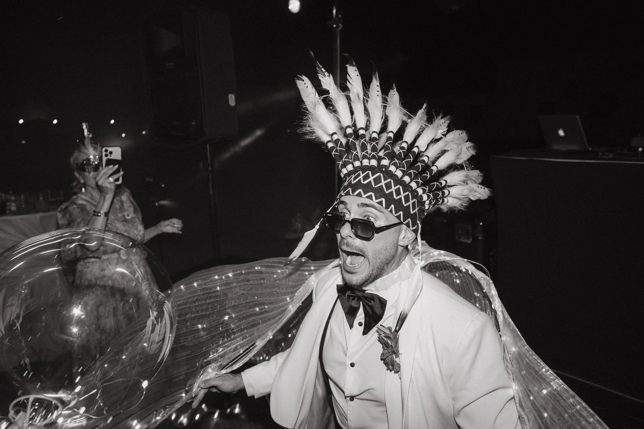 Un hombre vestido con un traje formal y un tocado con plumas, con gafas oscuras, en una expresión de sorpresa o emoción, en un ambiente oscuro con decoraciones y globos.