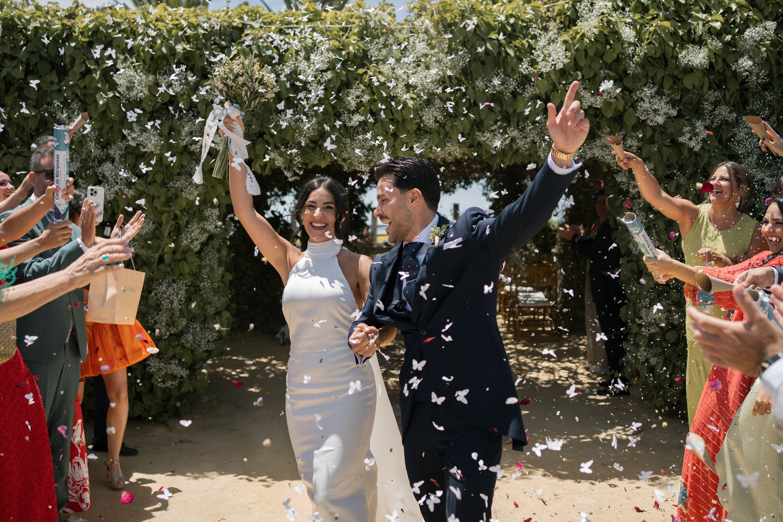Un par de recién casados celebrando su boda con amigos, rodeados de confeti y pétalos de flores en un entorno al aire libre.