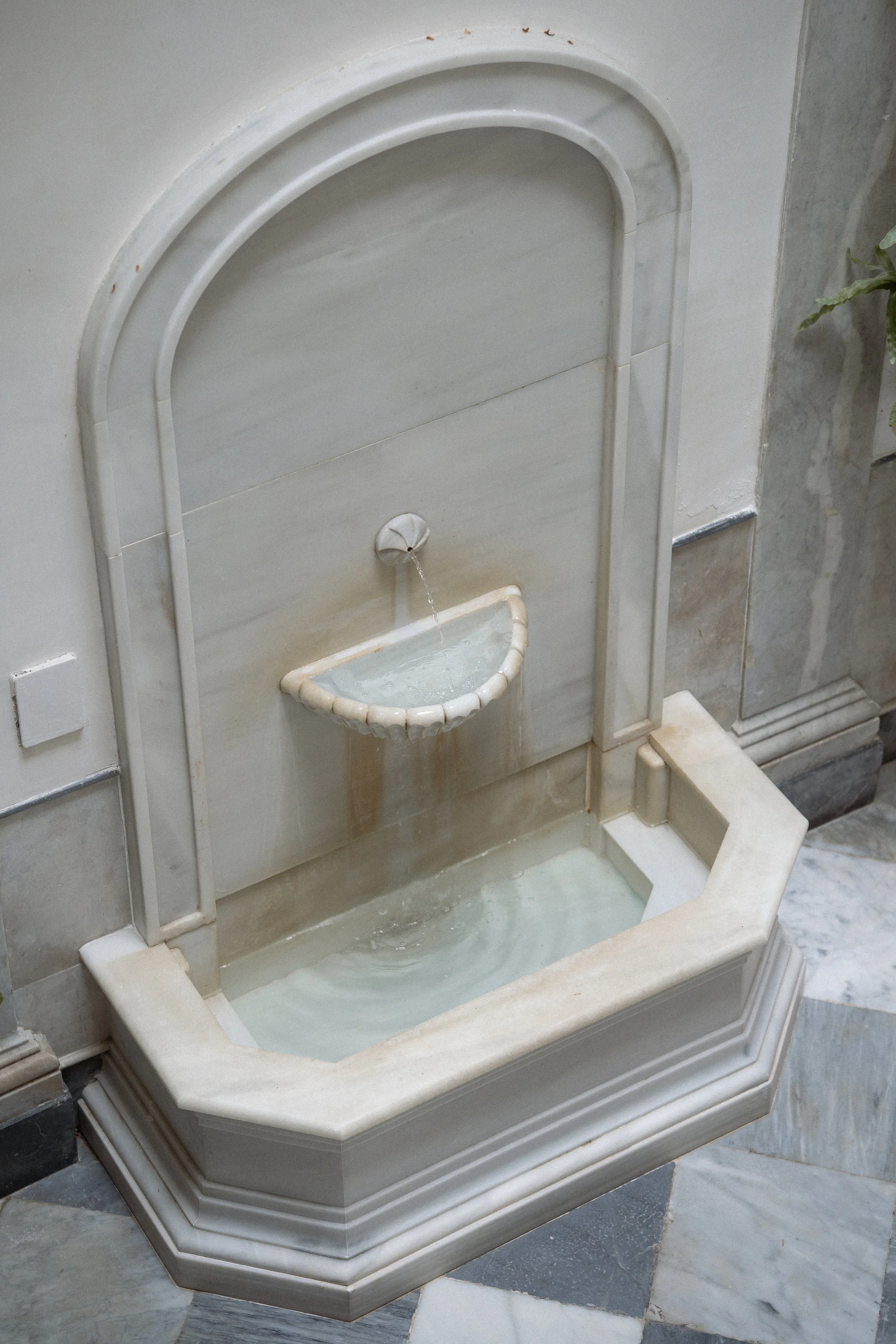 Fuente de agua decorativa de mármol con un pequeño caño y una cacerola colgante que vierte agua en un recipiente aprismado en la base, en un acabado en mármol y piedra. 