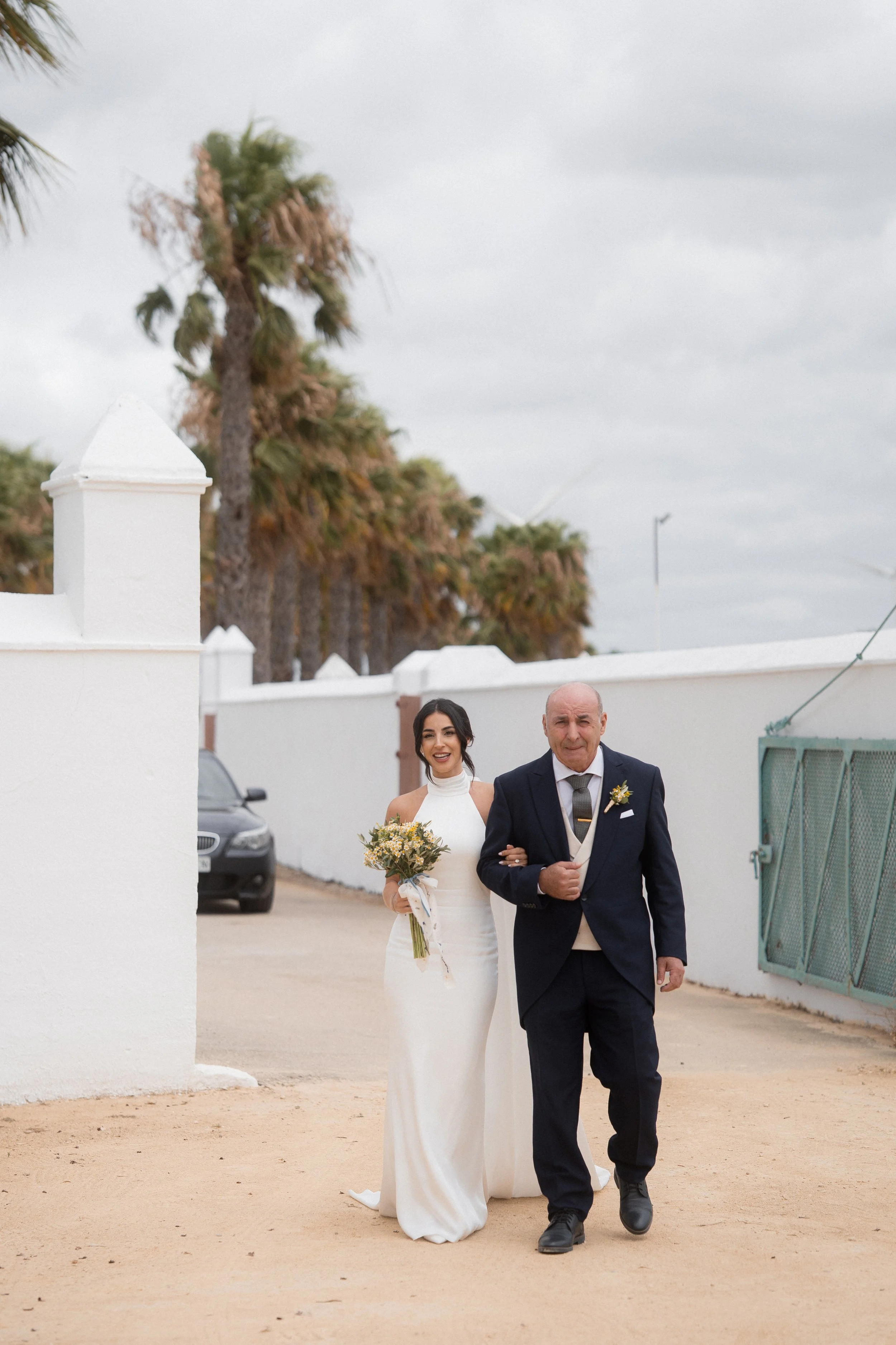 Mujer en vestido de novia con ramo de flores caminando con su padre en traje en un área exterior con palmeras y muro blanco.