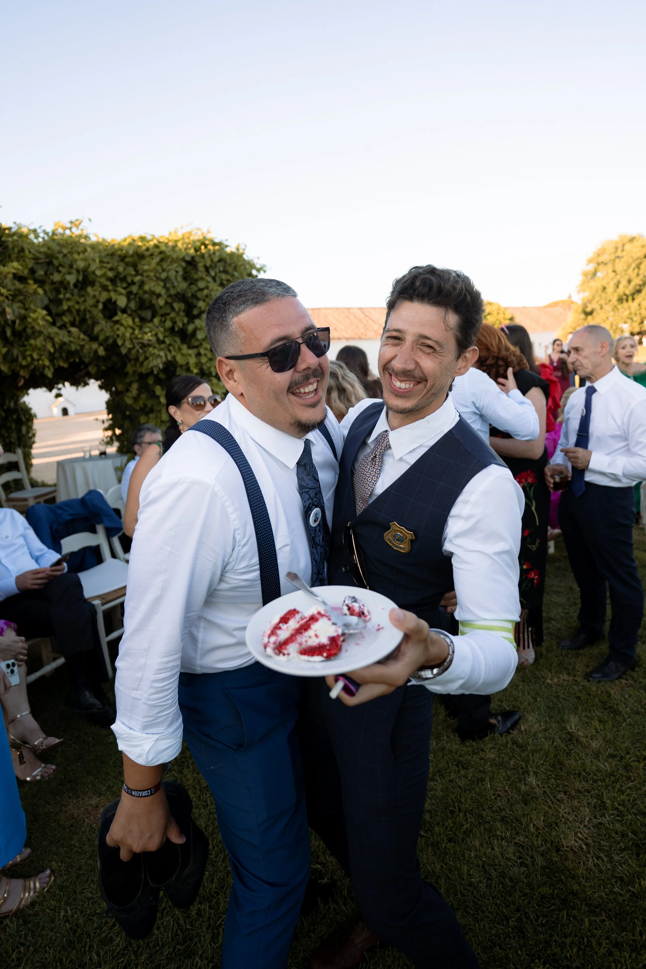 Dos hombres en ropa formal, uno con gafas de sol, sonriendo y abrazándose en una fiesta al aire libre, con una porción de pastel en un plato en manos de uno de ellos