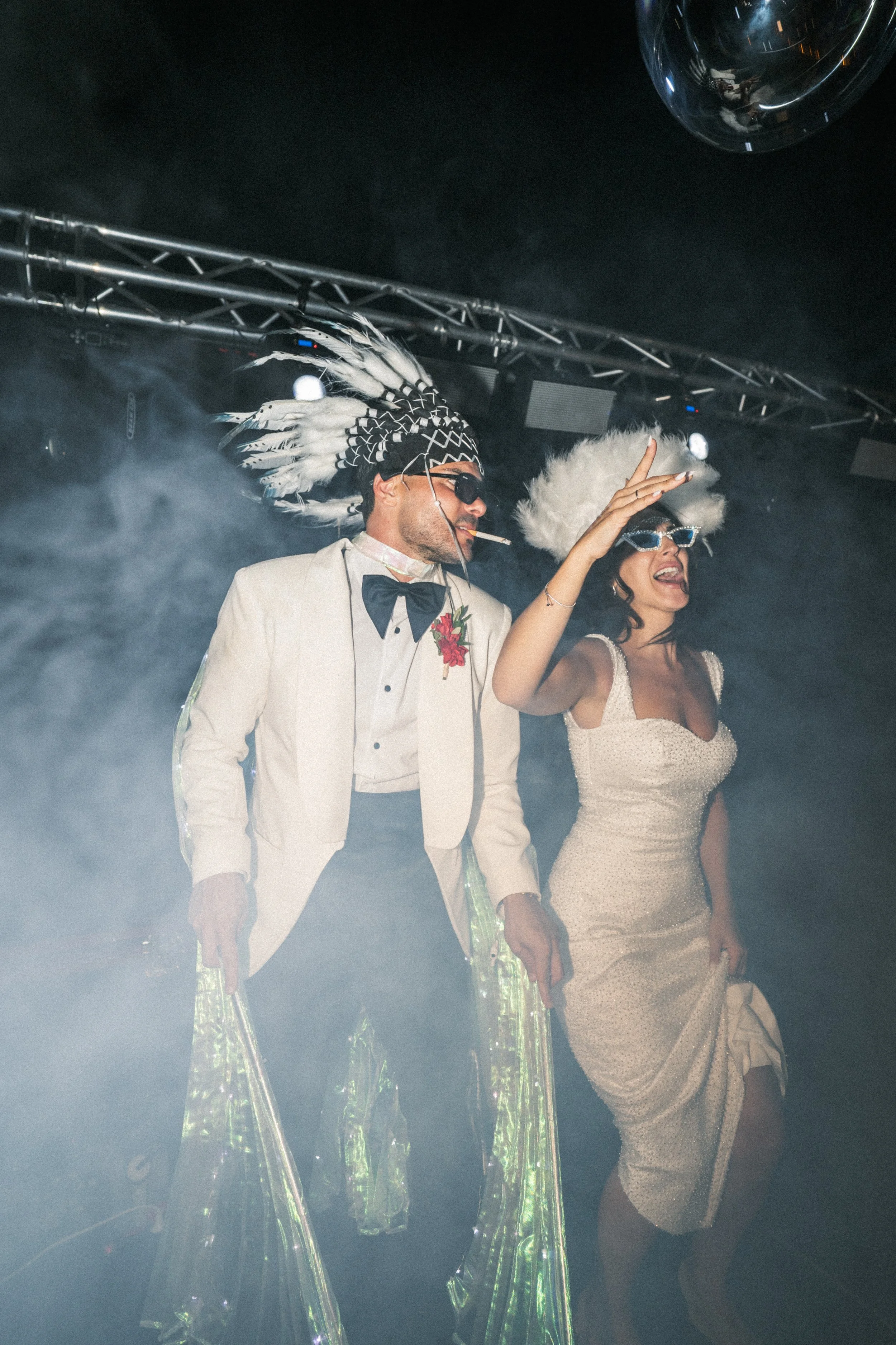 Persona con vestimenta formal dorada y accesorios en un evento, con una cabeza con plumas y gafas oscuras, y otra persona con un vestido y una peluca de peluche, en un ambiente con humo y luces de escenario.