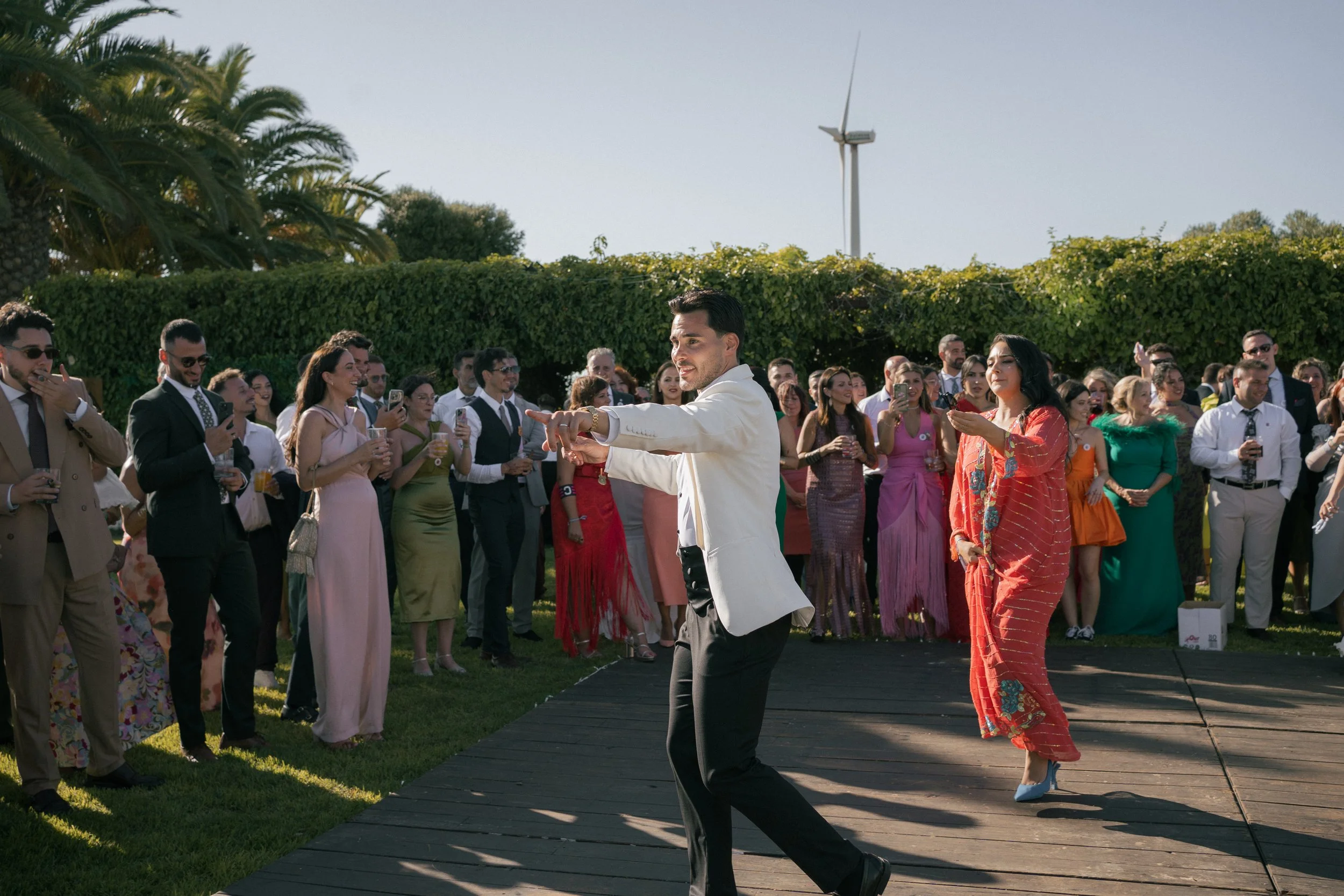 Pareja bailando en una ceremonia al aire libre con invitados observando y tomando fotos, en un entorno con árboles y un aerogenerador en el fondo.