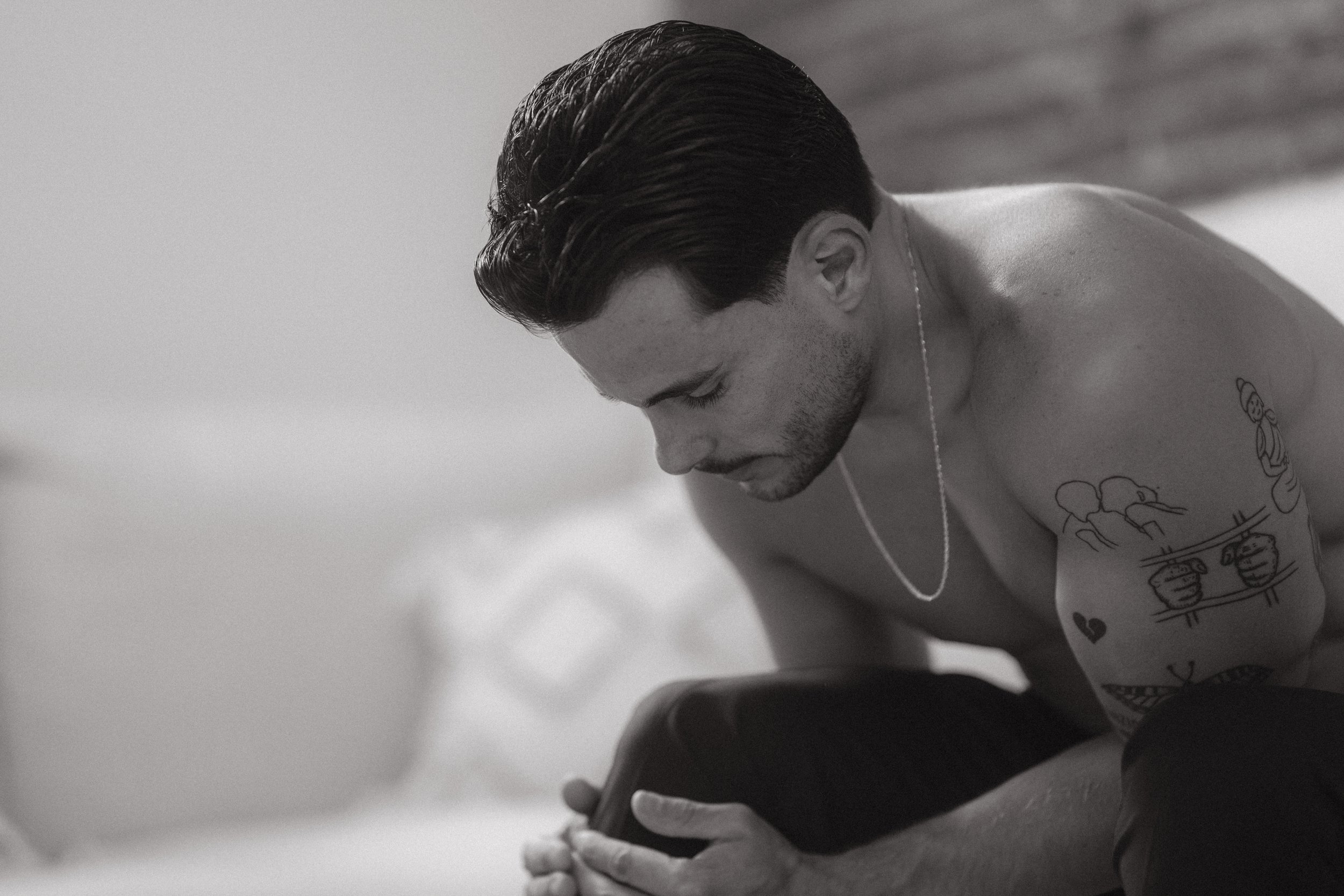 Hombre sin camisa con tatuajes, con cabello oscuro y peinado hacia atrás, mira hacia abajo mientras sostiene un teléfono móvil en sus manos, en un entorno interior.