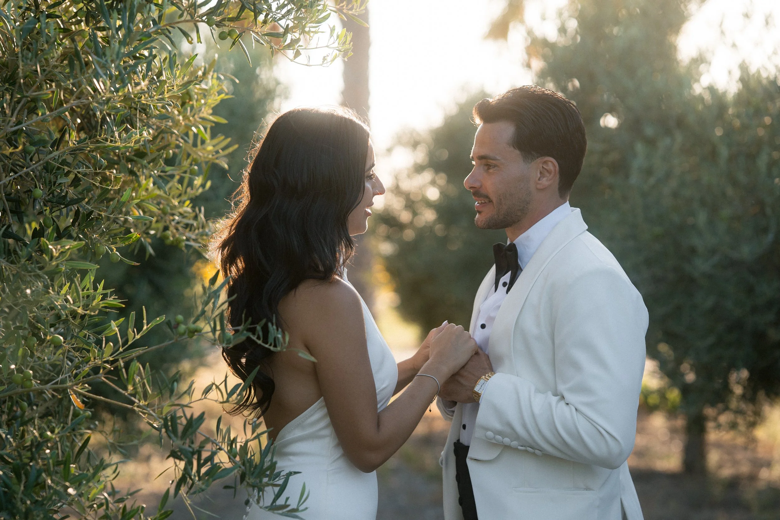 Pareja sonriente en una ceremonia al aire libre, la mujer con vestido blanco y el hombre con esmoquin blanco, sosteniéndose de las manos frente a árboles y al atardecer.