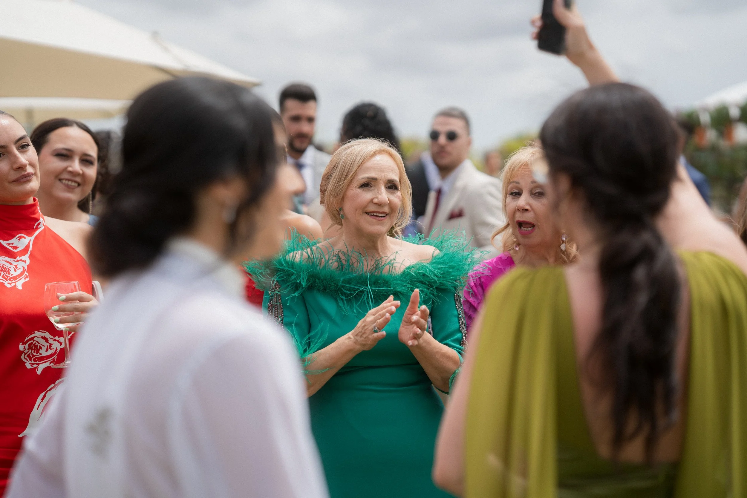 Grupo de personas conversando en un evento al aire libre, una mujer mayor en el centro usa un vestido verde con plumas en el cuello y está sonriendo, rodeada de otras mujeres y hombres que parecen disfrutar la ocasión.