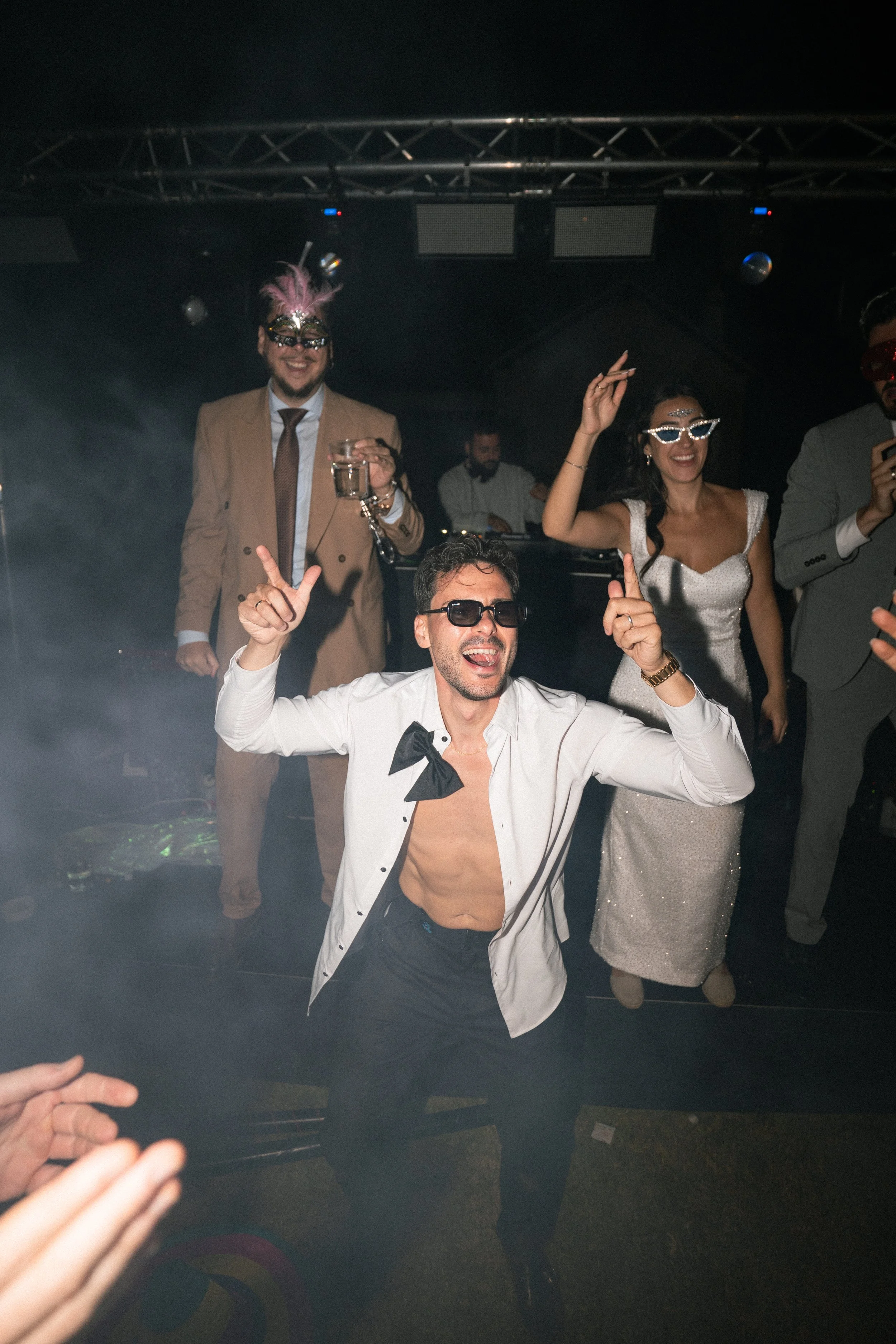 Personas bailando en una boda y disfrutando con disfraces y gafas de sol en un ambiente de fiesta con DJ en el fondo.