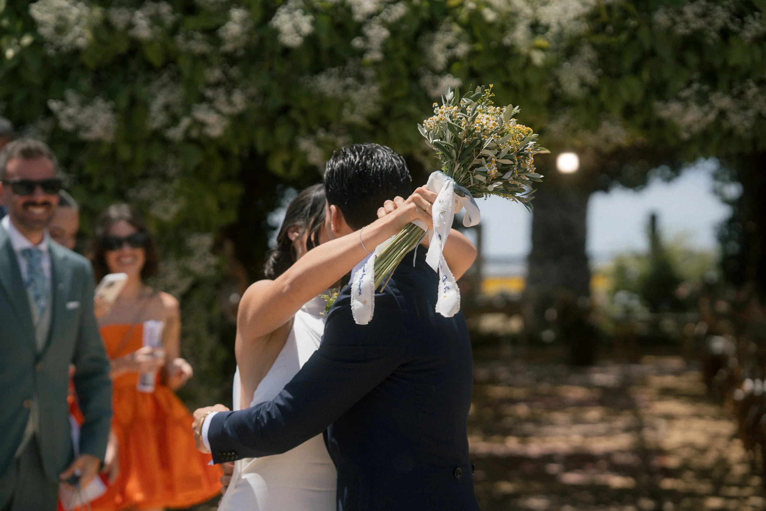 Una pareja de recién casados se abrazan y se besan, ella le pone un ramo de flores en la cabeza, en un entorno de árboles con invitados sonriendo y tomando fotos.