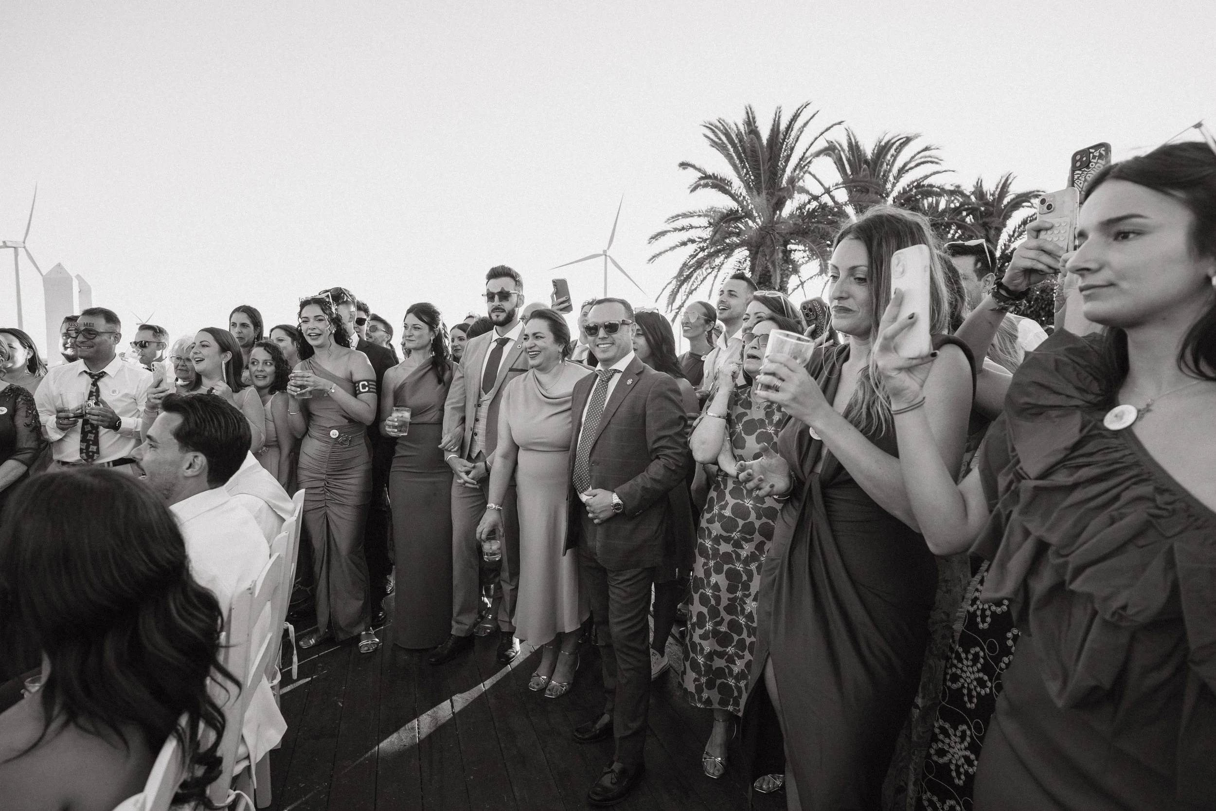 Un grupo de personas vestidas formalmente en un evento al aire libre, con palmeras y aerogeneradores en el fondo, algunos tomando fotos y otros observando.