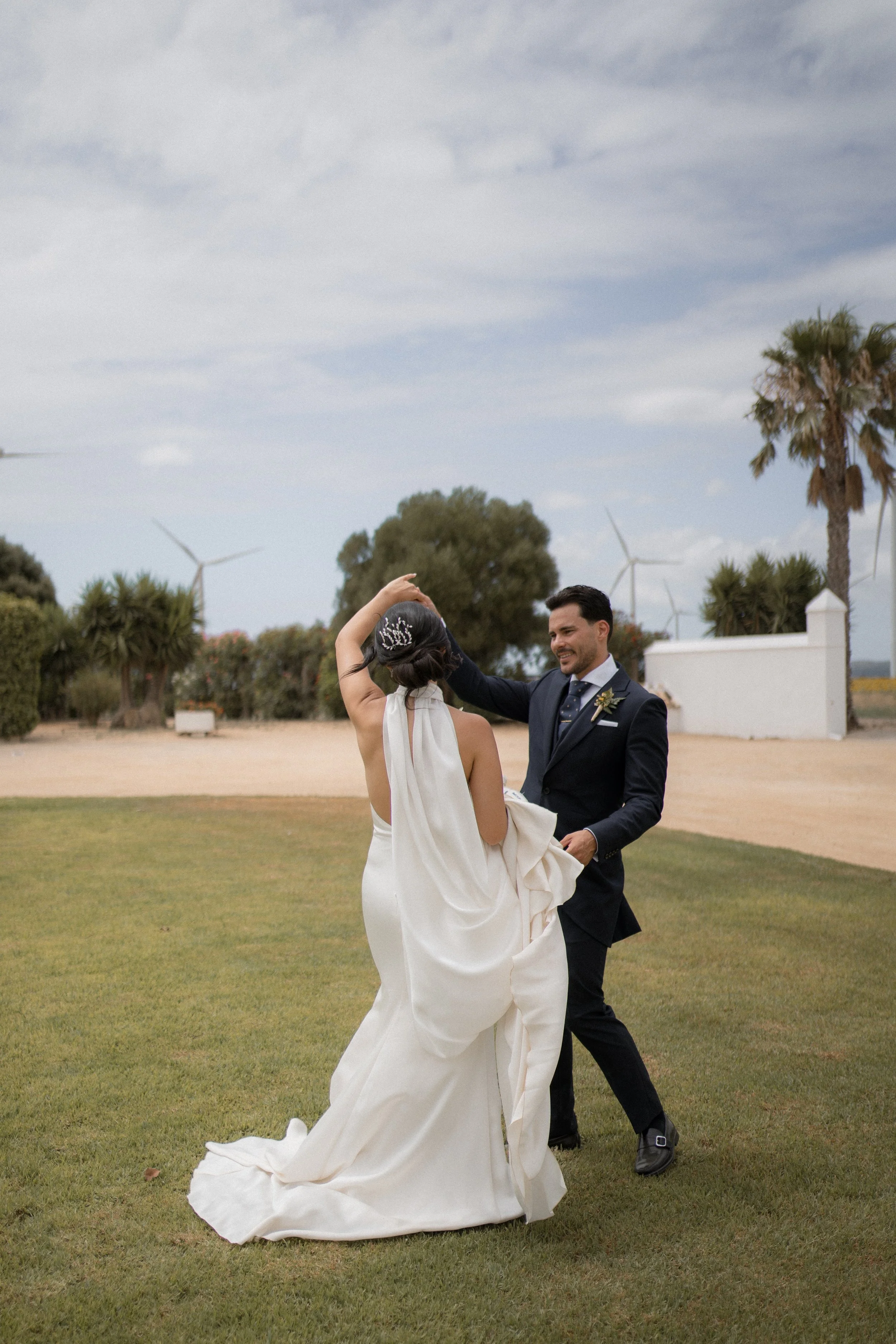 Una pareja bailando en un campo verde, con árboles y turbinas eólicas al fondo, en un día nublado.