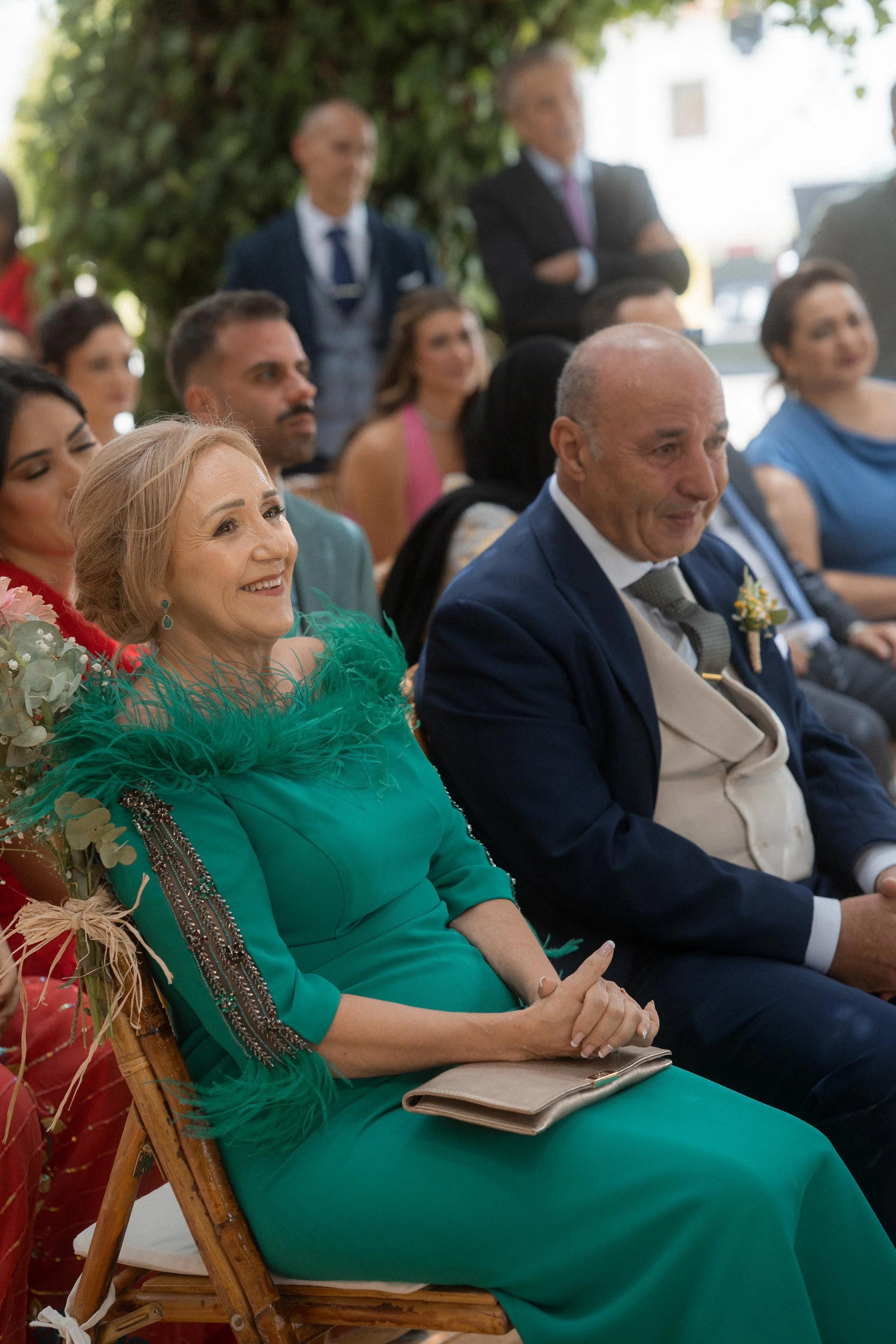 Hombres y mujeres en una ceremonia, algunos sonriendo, con vestimenta formal, en un ambiente al aire libre con árboles.