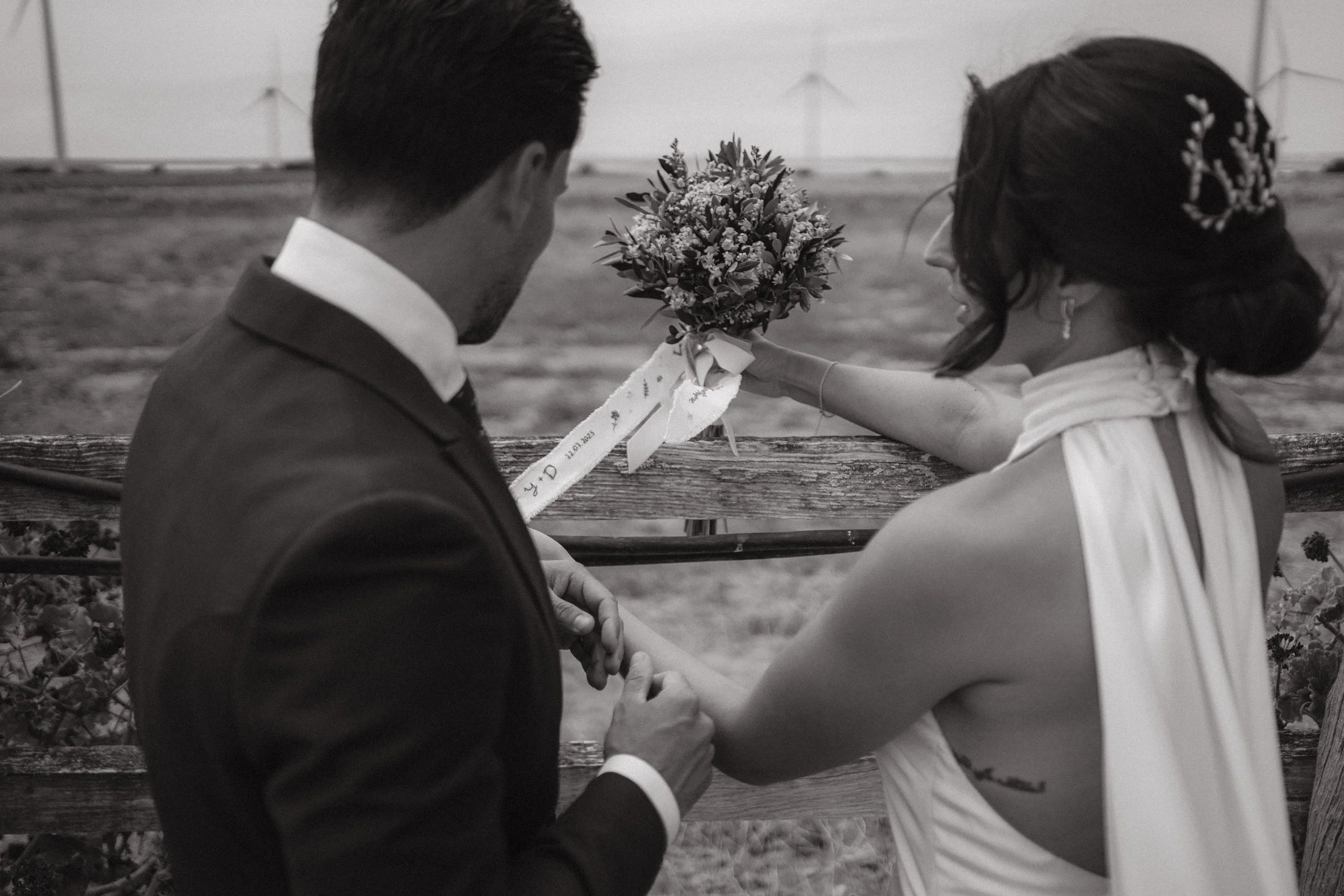 Una pareja de novios en una boda al aire libre, ella le entrega un ramo de flores al novio, en un entorno rural con un campo y estructuras metálicas en el fondo.