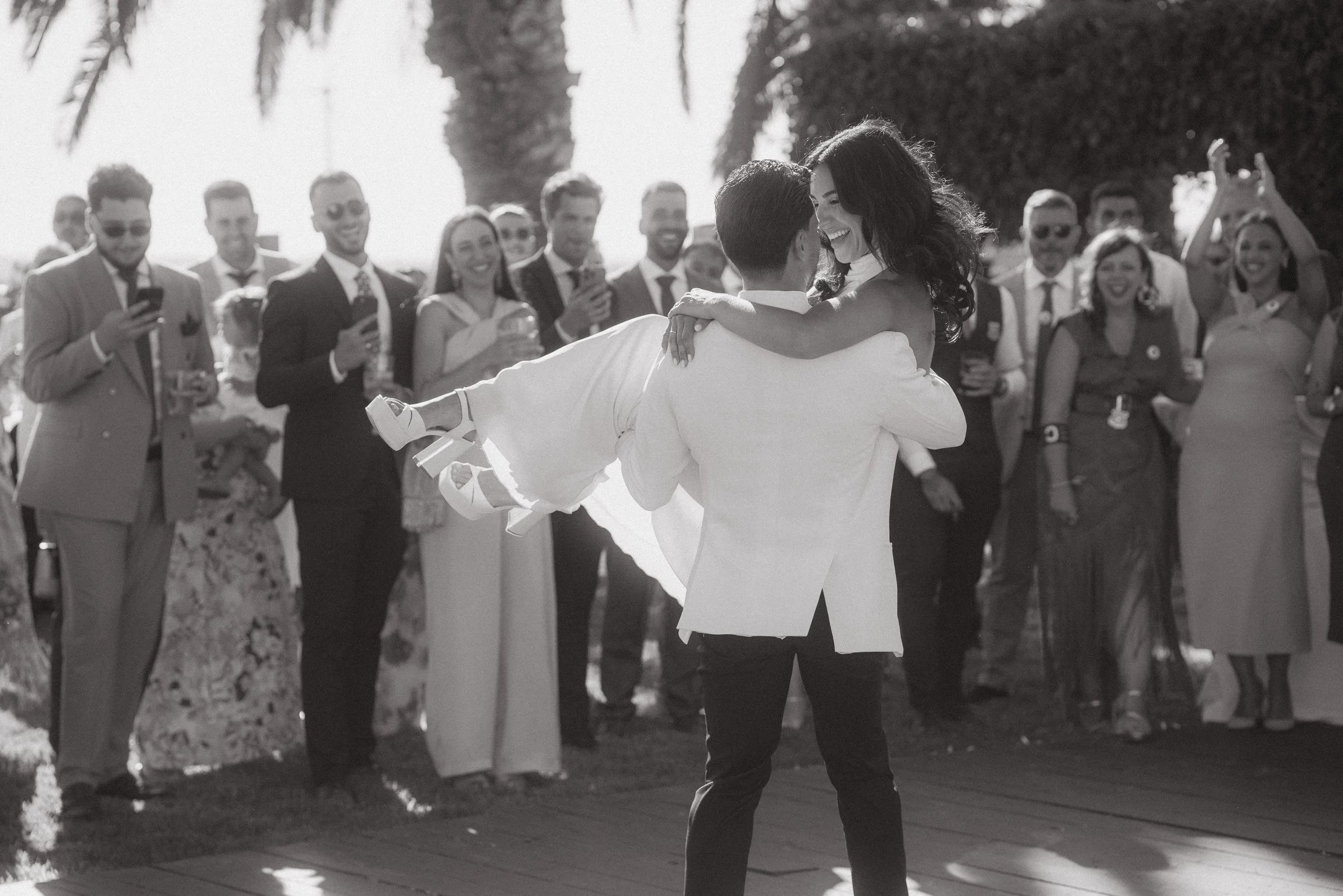 Una pareja bailando en una boda, rodeada de amigos y familiares con sonrisas, en un entorno al aire libre con palmeras.