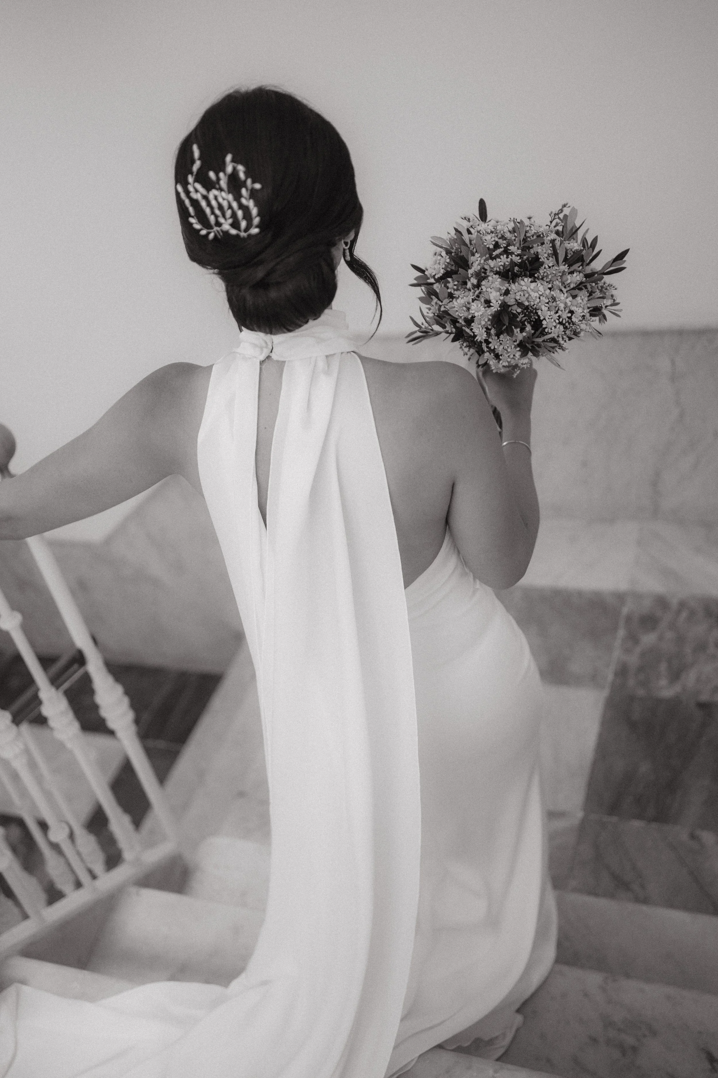 Mujer con vestido de novia y peinado elegante, sosteniendo un ramo de flores en una escalera de interior.