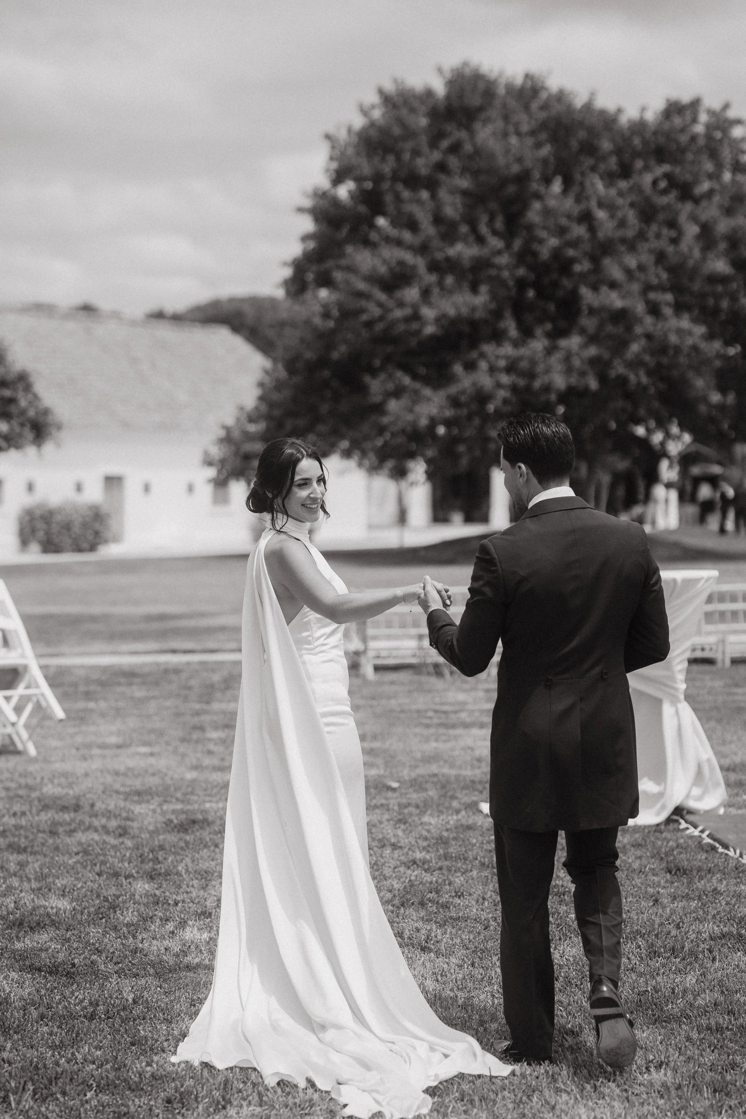 Una pareja en atuendo de boda bailando en un lugar al aire libre con árboles y sillas blancas, en blanco y negro.