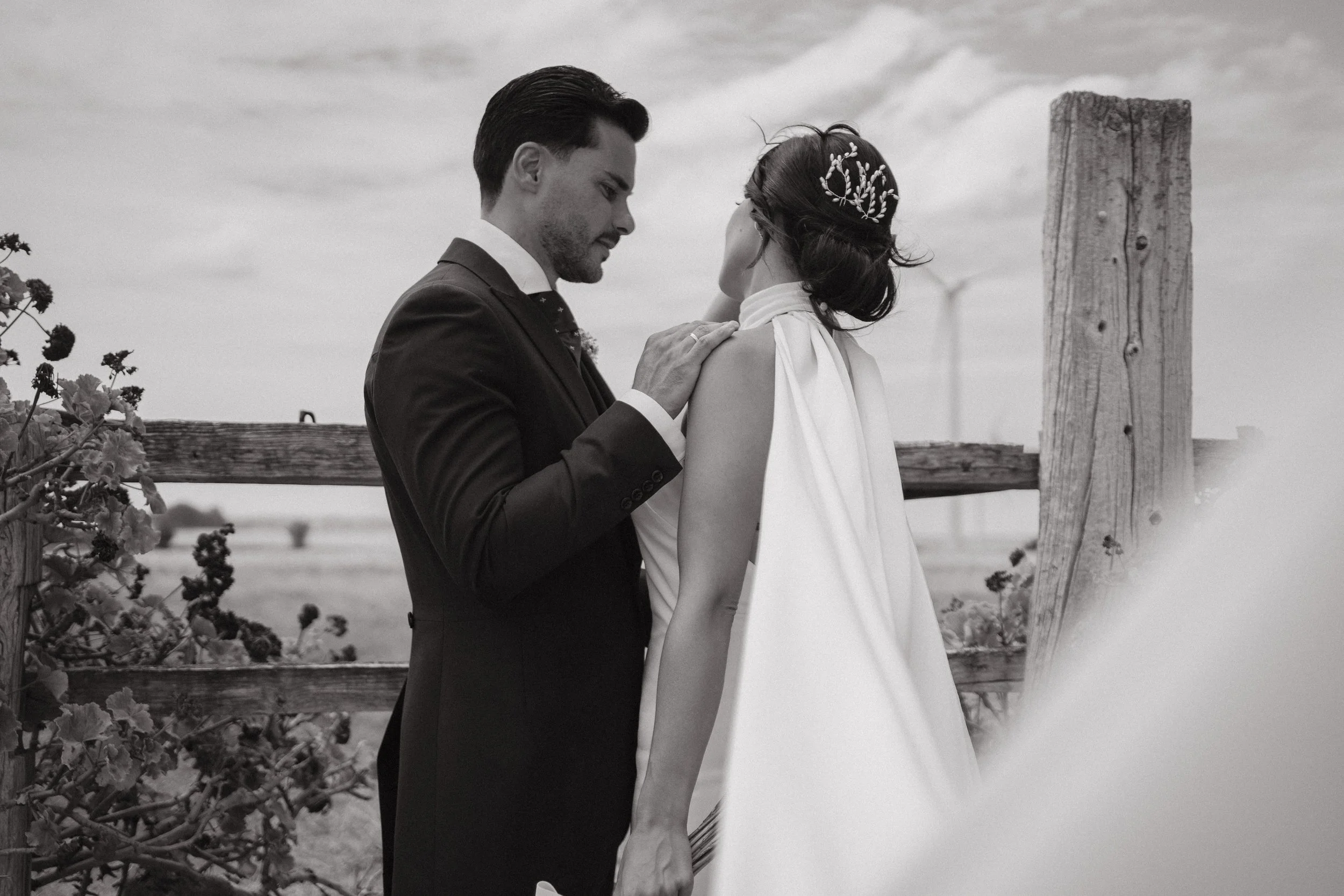 Una pareja en boda en un escenario al aire libre con fondo de cielo y molinos de viento, en un estilo en blanco y negro.