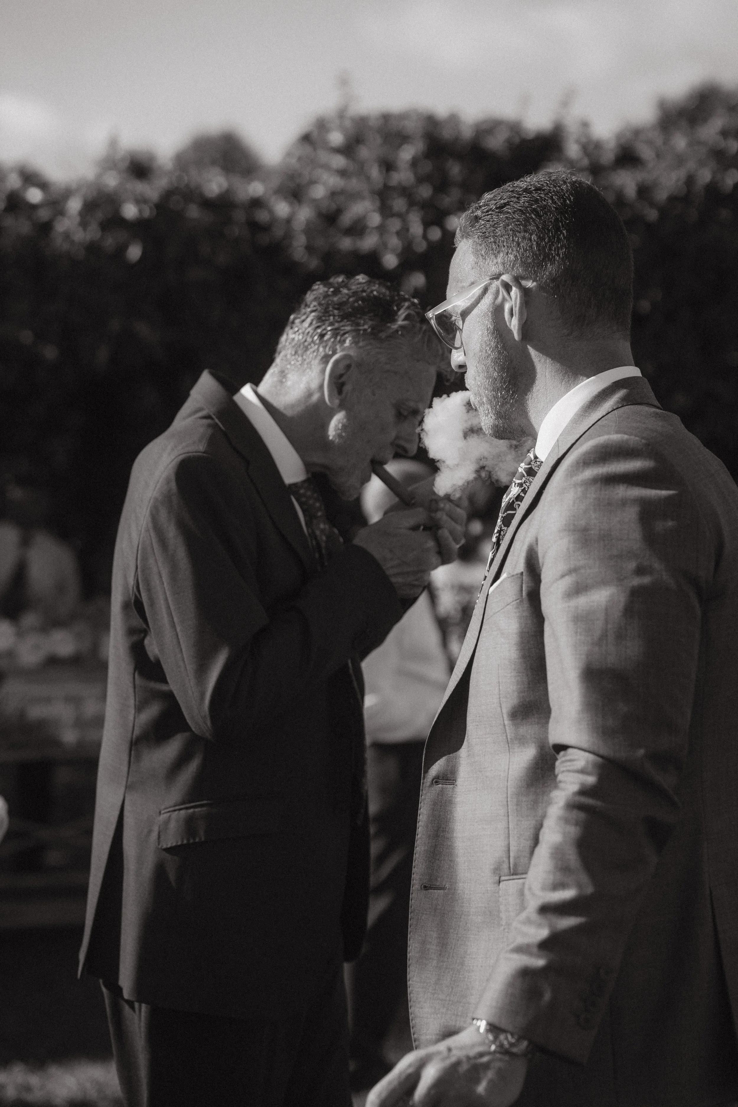 Dos hombres vestidos con trajes de etiqueta, uno de ellos fumando un cigarrillo. En un entorno exterior con árboles en el fondo, la imagen en blanco y negro captura un momento de interacción entre ellos.