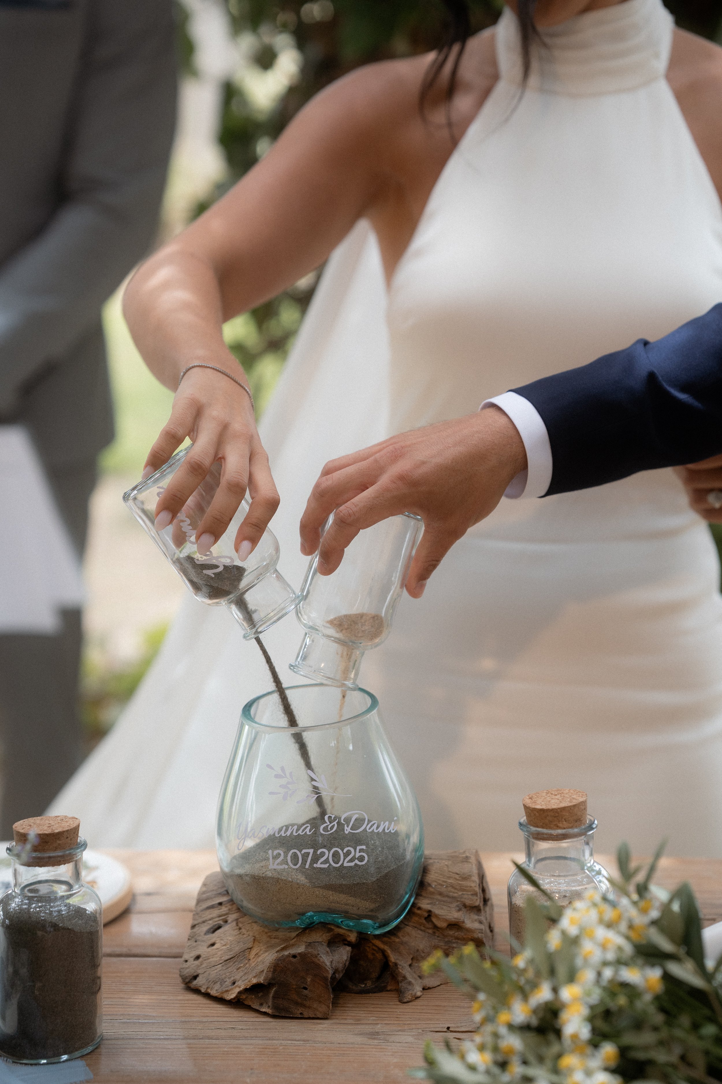 Una pareja casándose en una ceremonia al aire libre, vertiendo arena en un recipiente decorativo que tiene los nombres 'Yasmina & Dani' y la fecha '12.07.2025', simbolizando su unión.