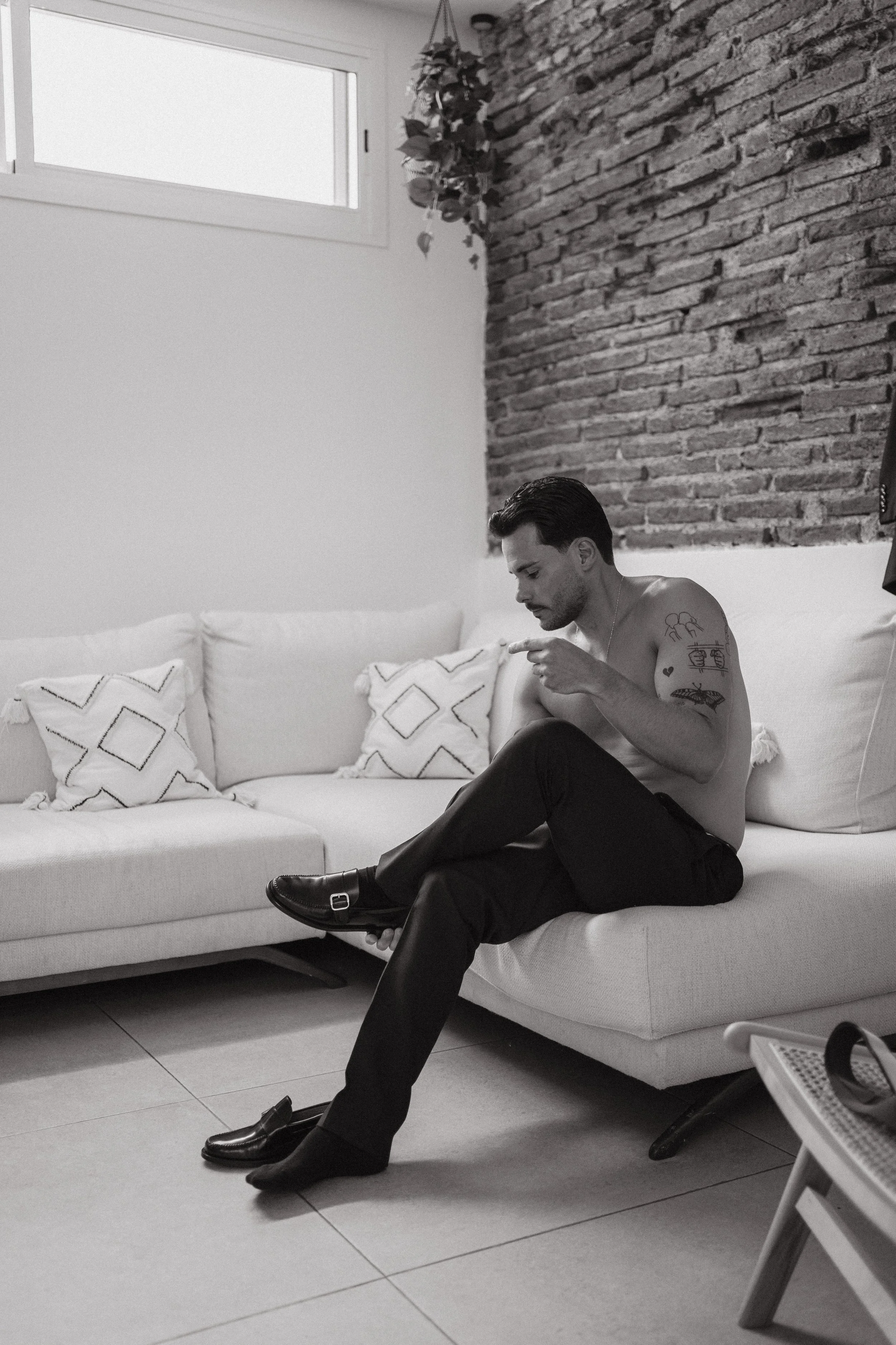 Un hombre sentado en un sofá blanco, sin camiseta, mirando su teléfono móvil, con zapatos quitados en el suelo.