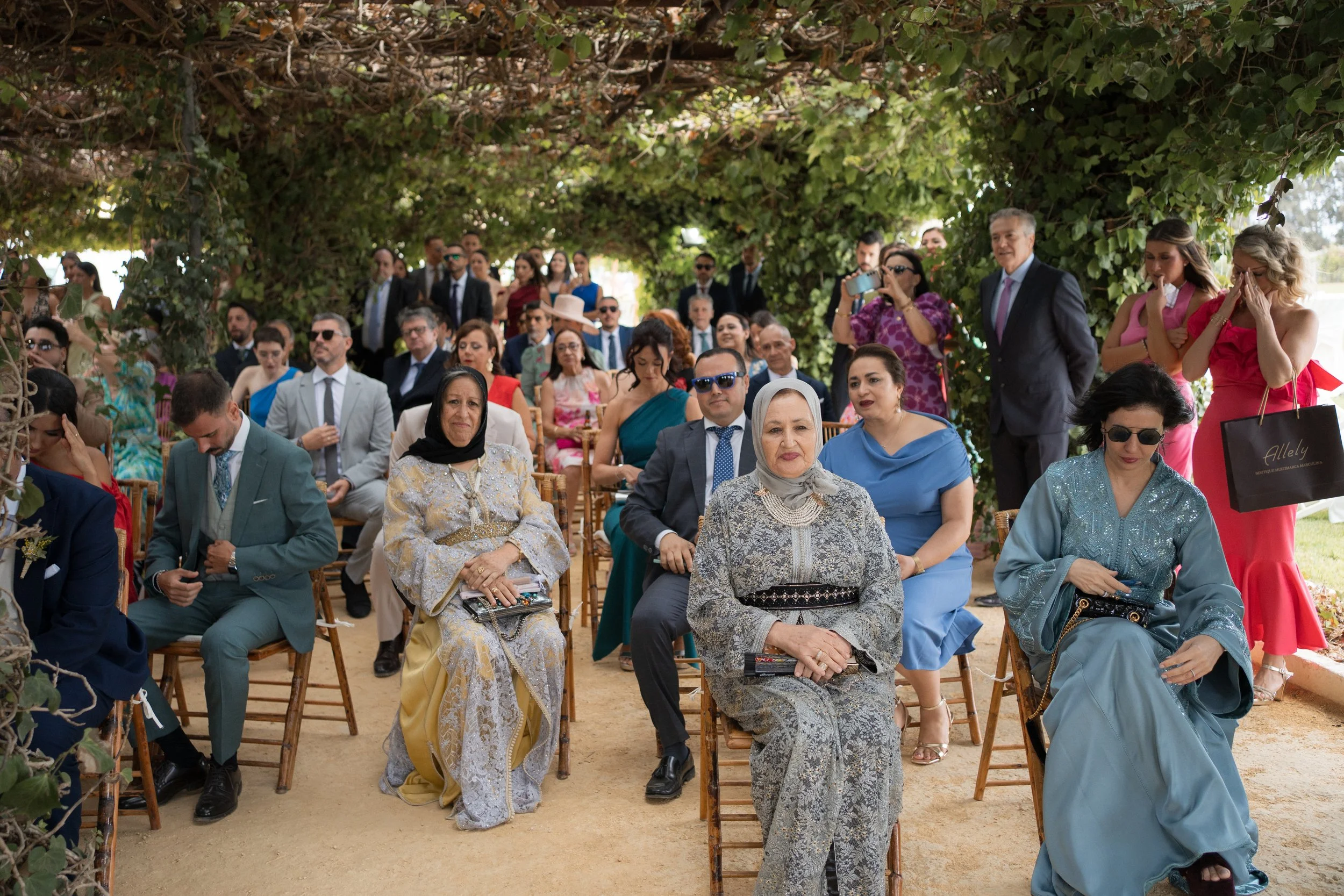Personas en una ceremonia al aire libre, sentadas y de pie, bajo un arco de follaje, rodeadas de vegetación y árboles.