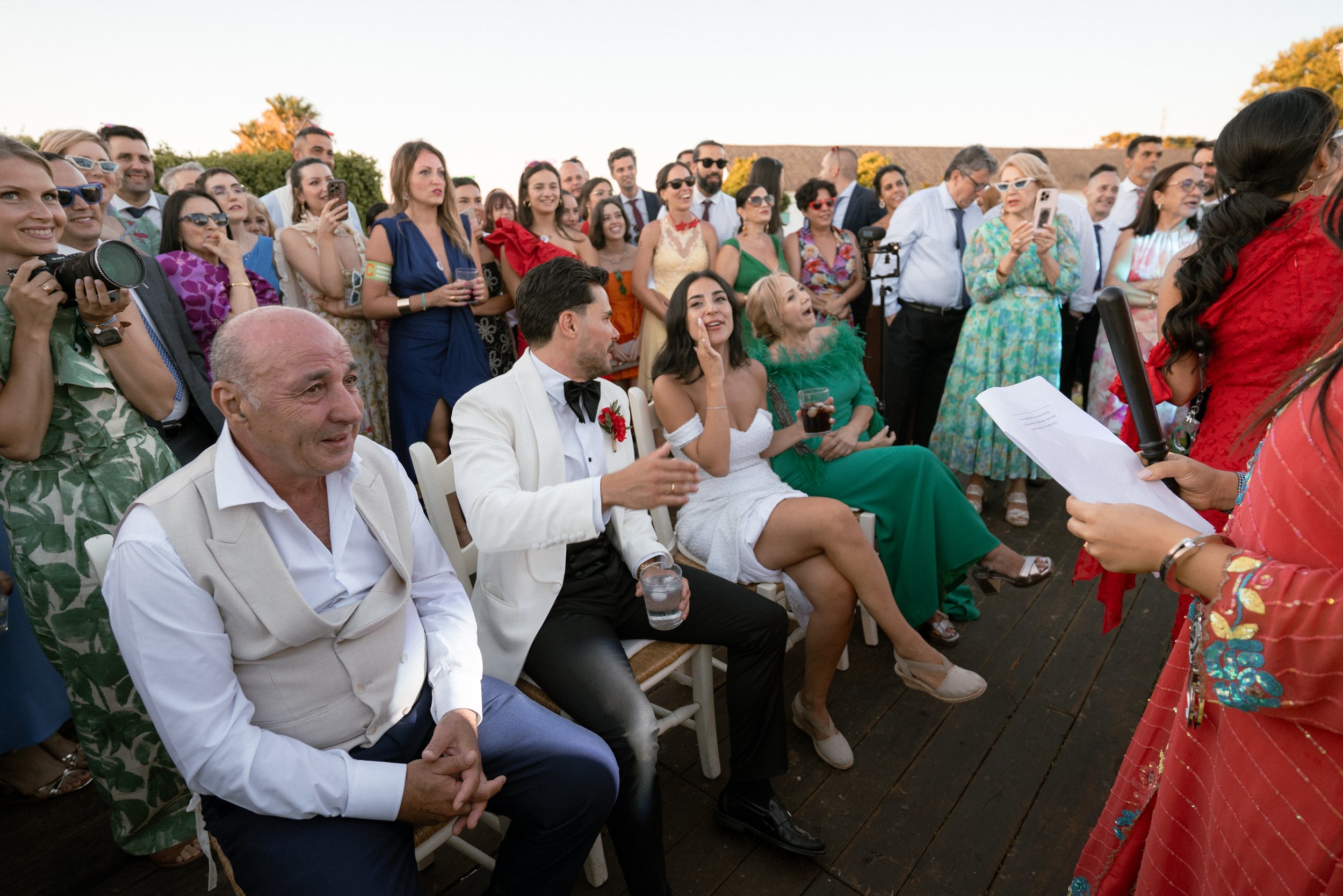 Reunión en una boda con invitados sentados y de pie, disfrutando el evento en un ambiente al aire libre, con una mujer con vestido blanco y un hombre con traje de etiqueta en primer plano.