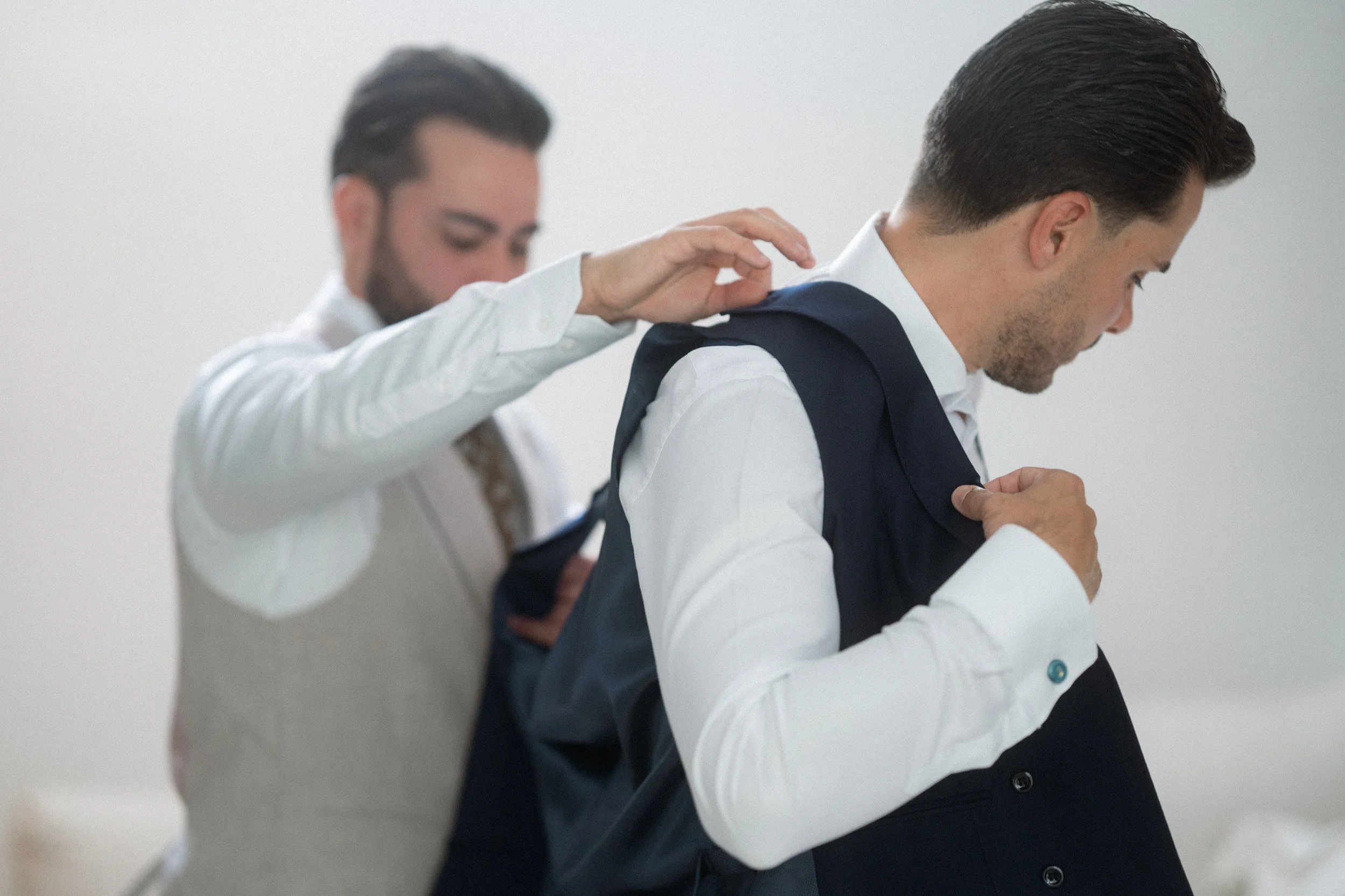 Dos hombres vestidos con camisas blancas, uno ayuda al otro a ponerse un saco oscuro.