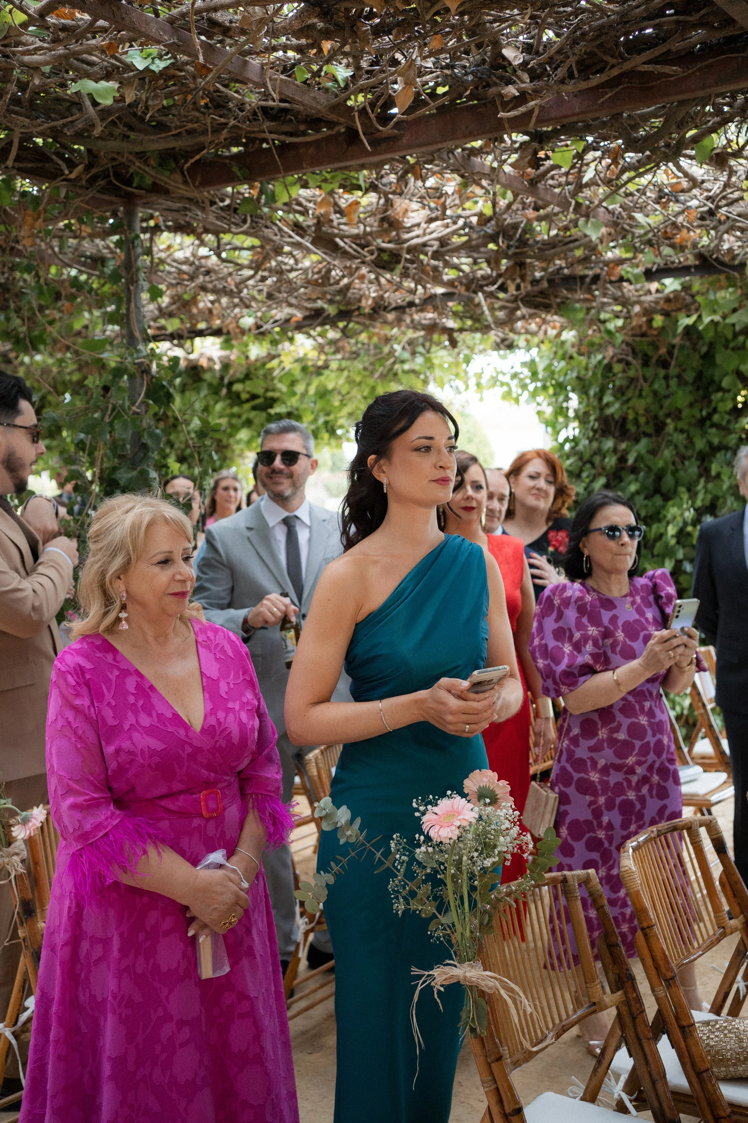 Grupo de personas vestido con ropa elegante en una ceremonia al aire libre bajo un techo de ramas con follaje. Algunas personas están usando gafas de sol y sosteniendo teléfonos móviles, mientras otras observan atentamente.