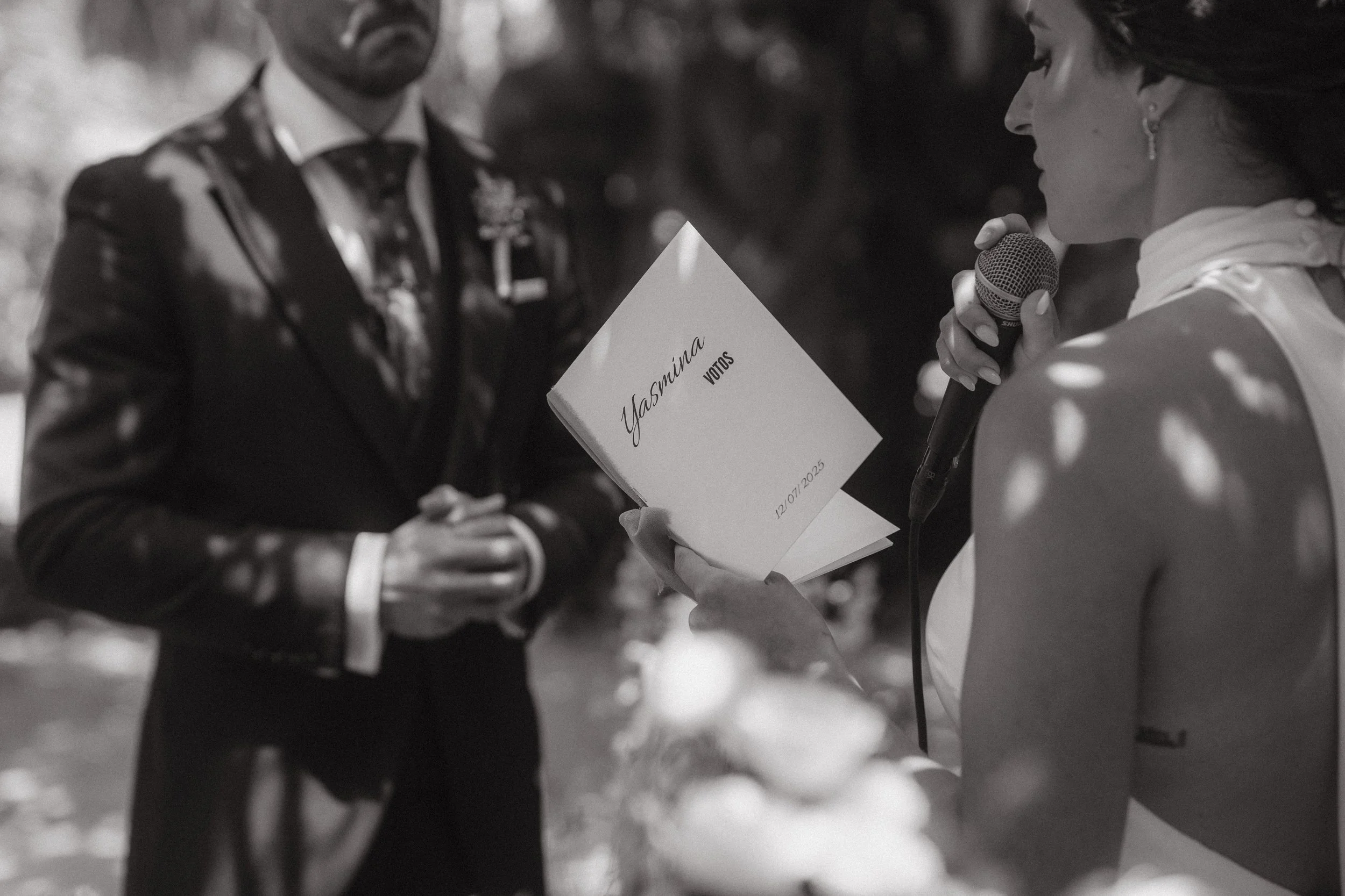 Mujer leyendo votos en una boda, con un libro que dice 'Yasmina Votos 12/07/2025', en presencia de un hombre en traje formal, en un entorno al aire libre.