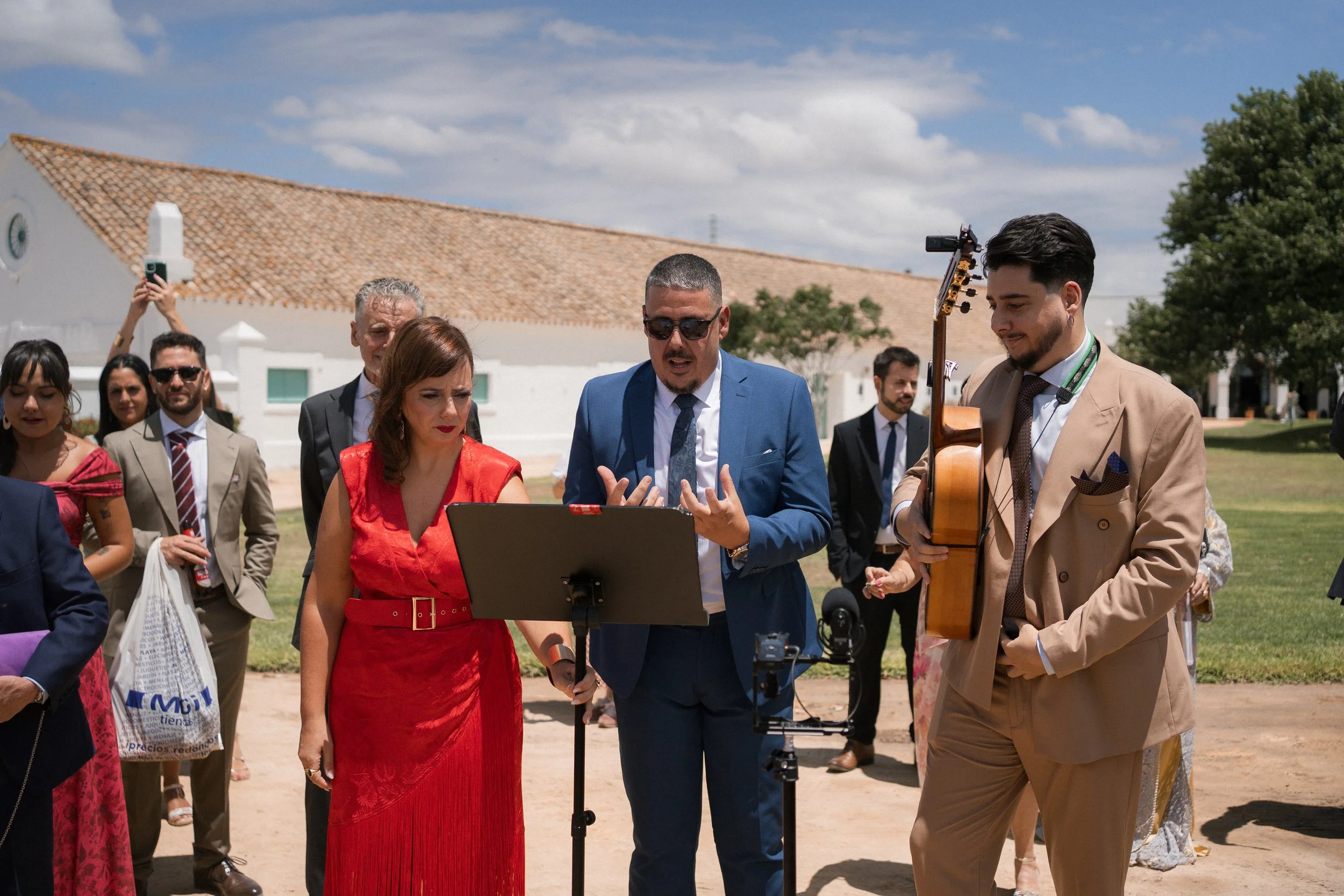 Grupo de personas en una ceremonia al aire libre, con una mujer en vestido rojo, dos hombres en traje azul y beige, uno tocando guitarra, y otros en el fondo.