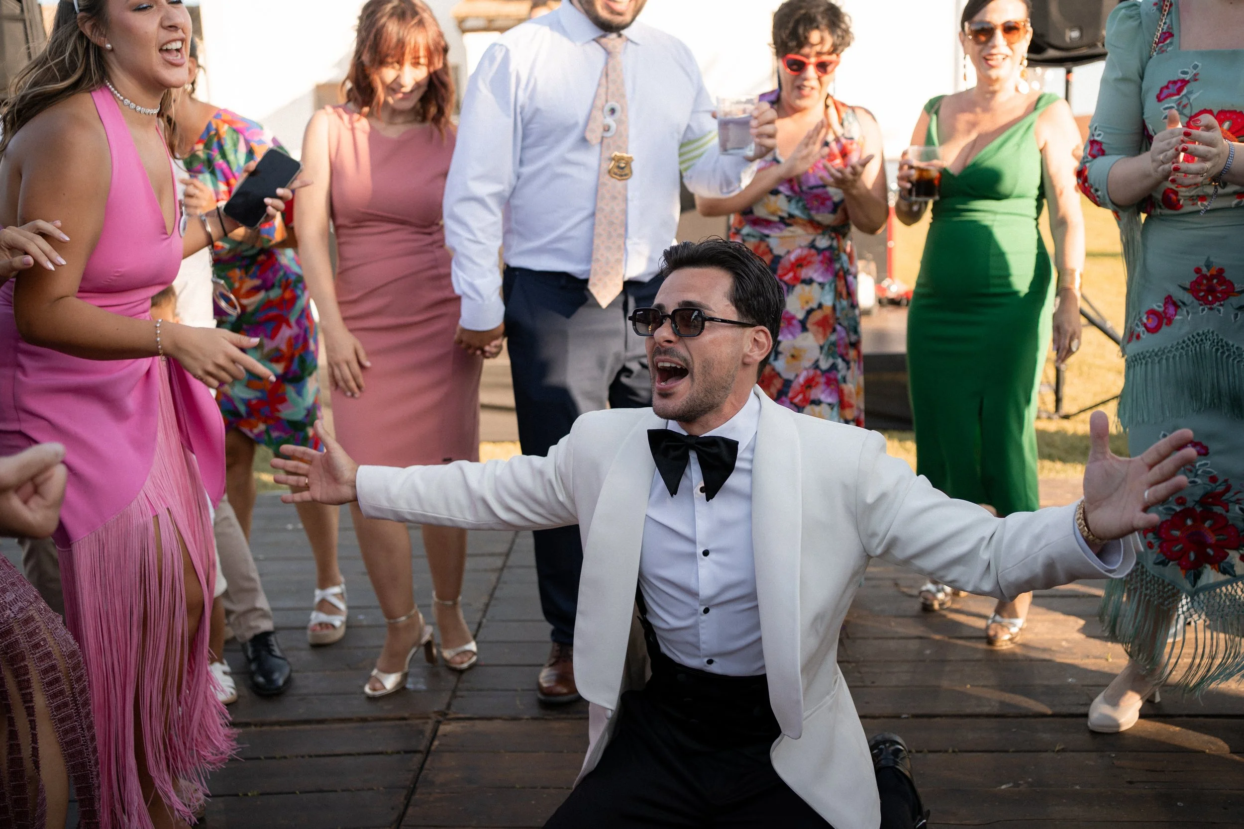 Un grupo de personas en una celebración al aire libre, con un hombre en el centro vestido con traje blanco y gafas oscuras, aparentemente bailando o celebrando con entusiasmo.
