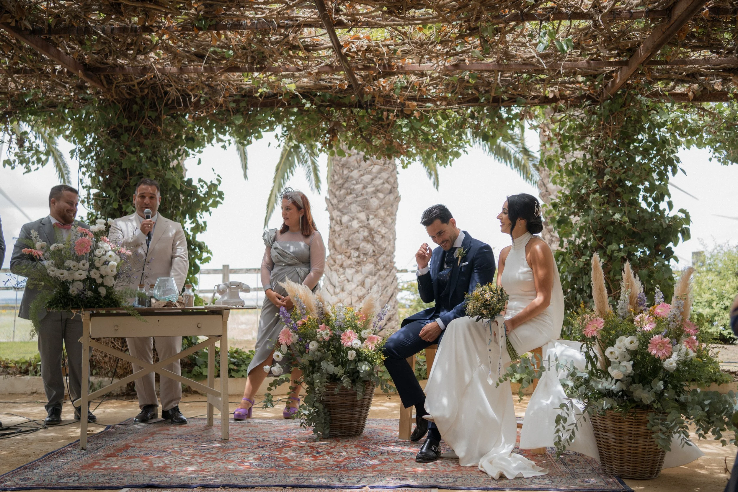 Pareja de novios en una boda al aire libre, rodeados de flores y vegetación, con un oficiante y dos testigos, bajo un arco decorado con plantas y flores.