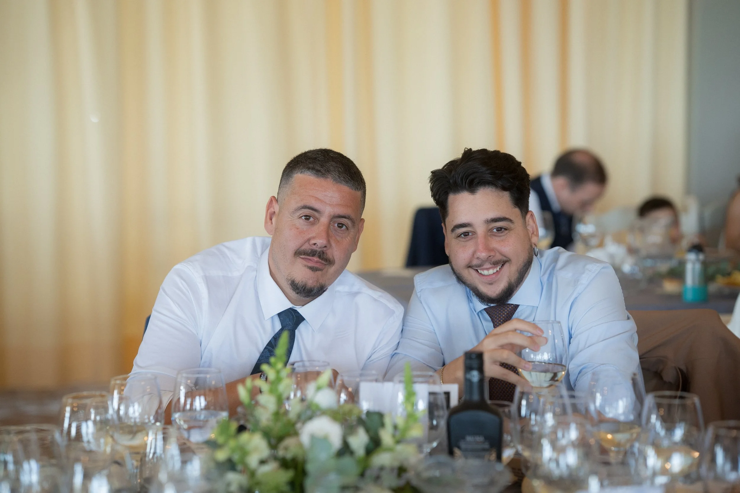 Los hermanos Pastrana en una boda, uno con barba y otro con barba y arete, sentados en una mesa con vino y en un ambiente de evento social.