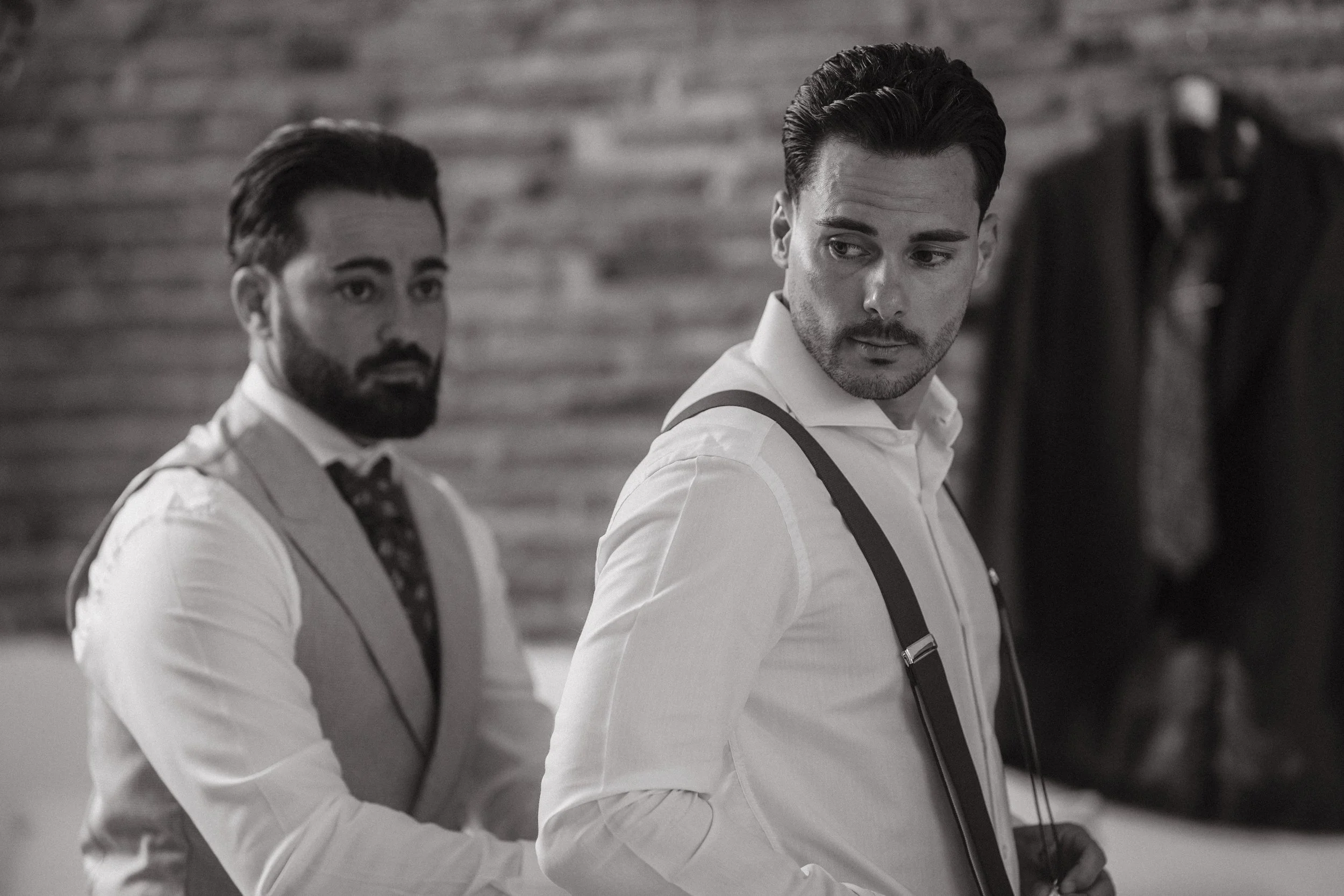 Dos hombres con ropa formal, uno con suspensores, en un ambiente con fondo de paredes de madera.