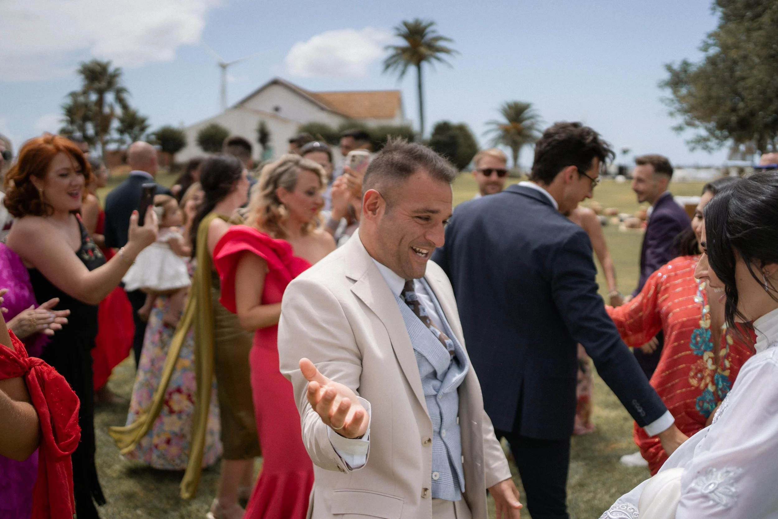 Jóvenes en una boda al aire libre, bailando y tomándose fotos, con árboles y casas en el fondo.