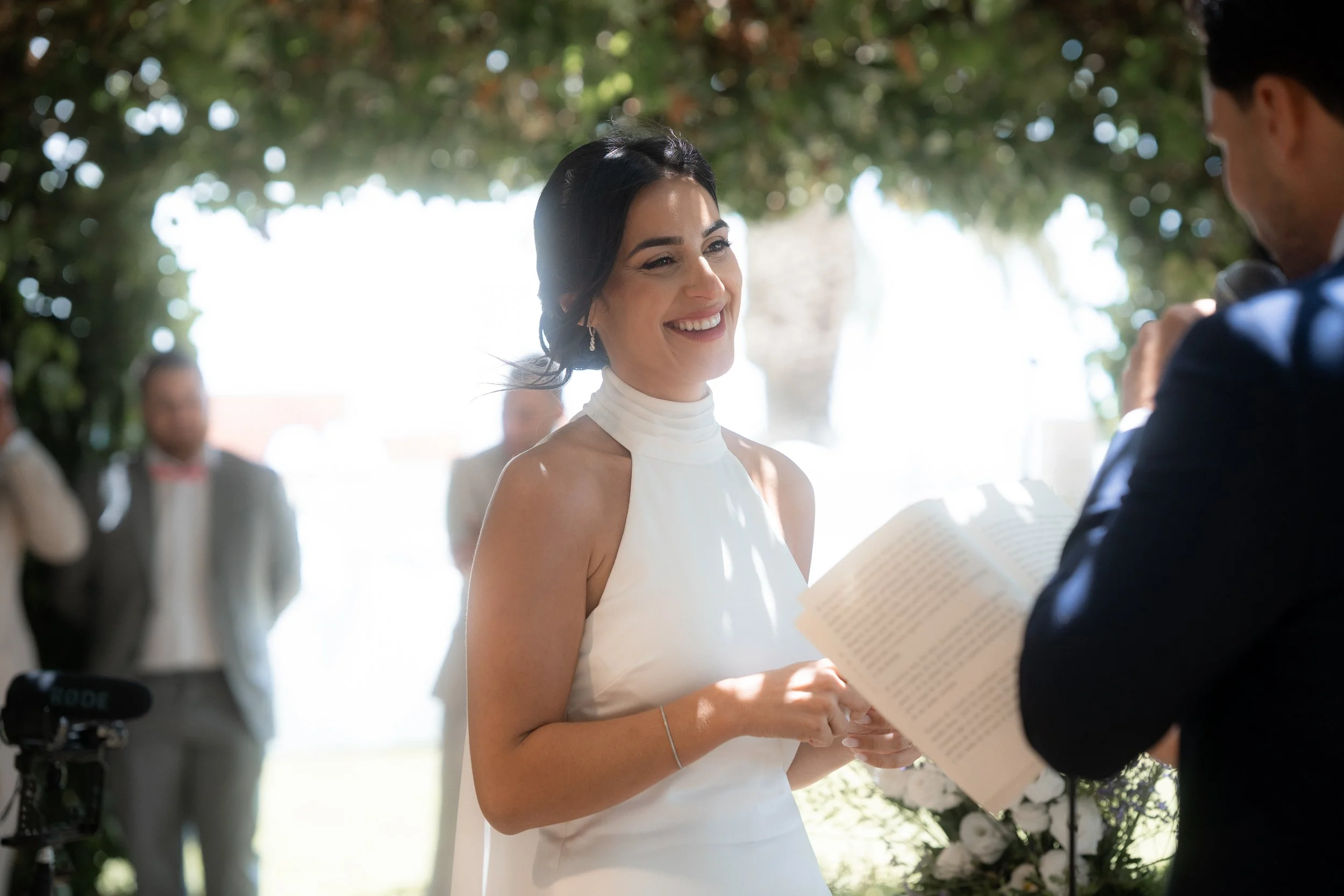 Mujer en vestido blanco sonriendo en una ceremonia al aire libre mientras intercambia votos o anillos con un hombre.