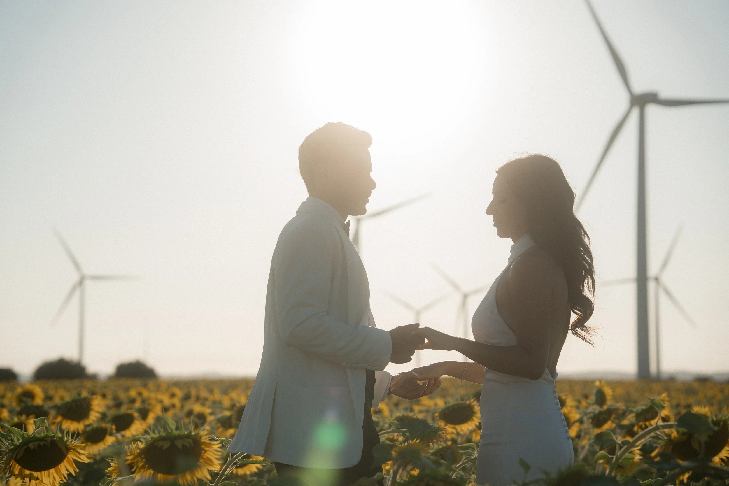 Una pareja sostiene una ceremonia en un campo de girasoles al atardecer, con aerogeneradores en el fondo.
