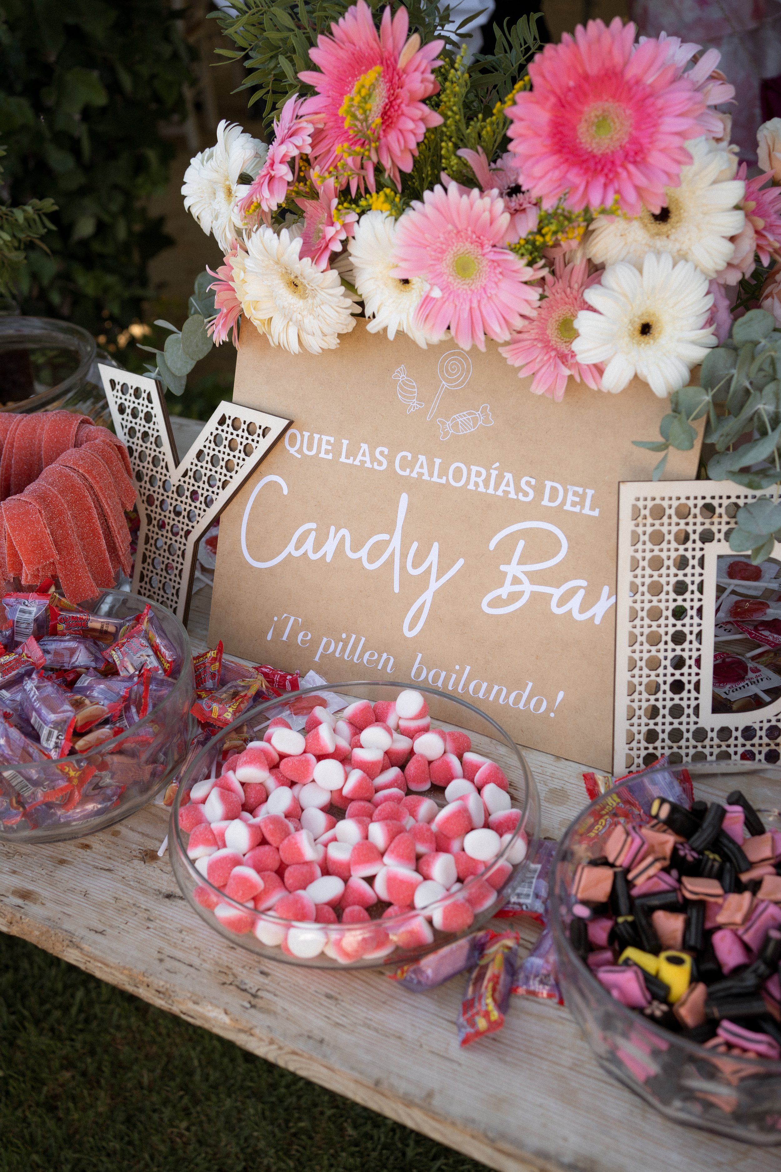 Mesa con dulces, una caja con caramelos de miel, una de gomitas, una de dulces en color rosa y negro. Decoraciones con flores y un cartel que dice 'Que las calorías del Candy Bar ¡Te pillen bailando!'