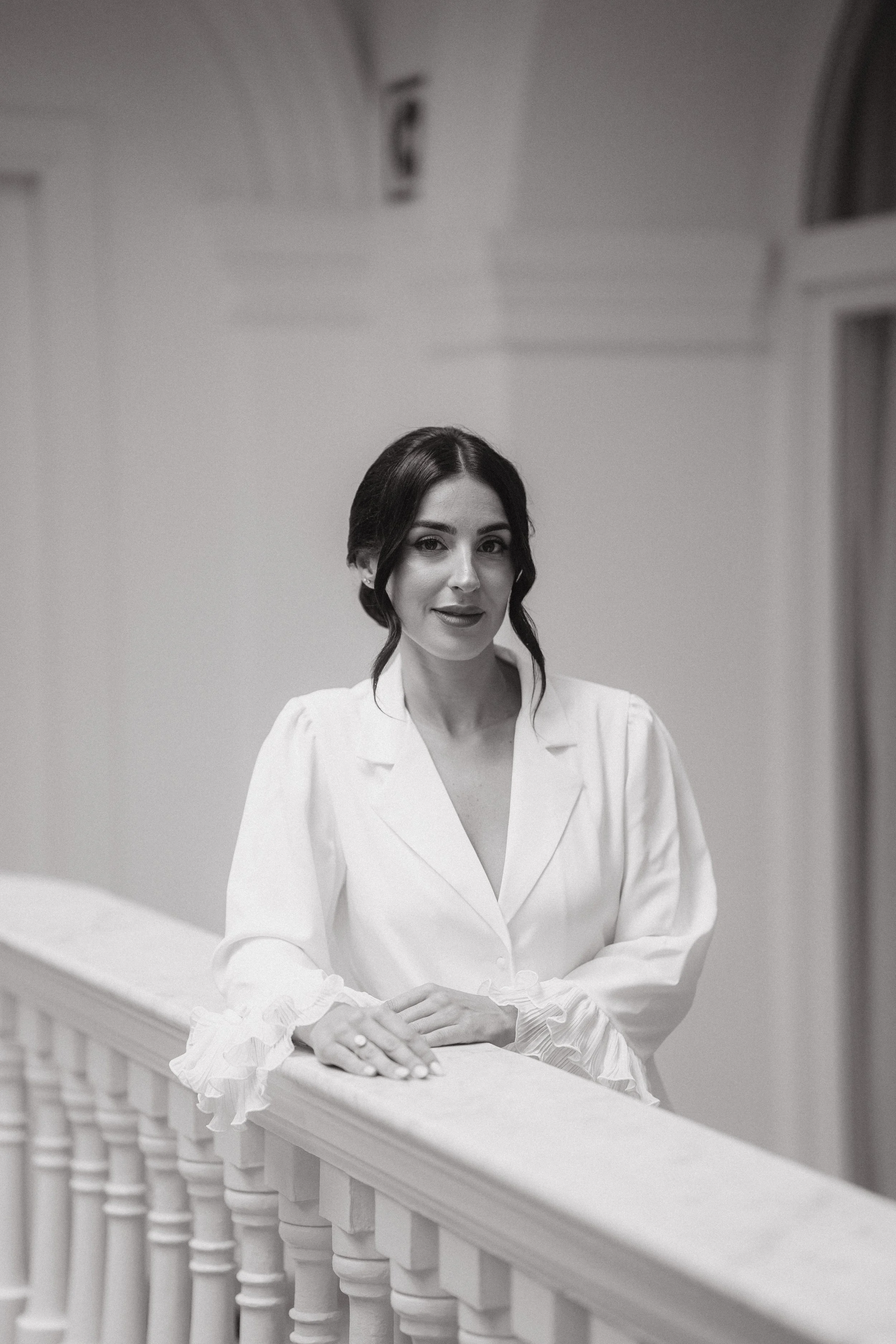 Mujer en una bata blanca, de cabello oscuro y peinado suelto, posando en un pasillo de edificio de estilo clásico con barandilla y ventanas en blanco y negro.