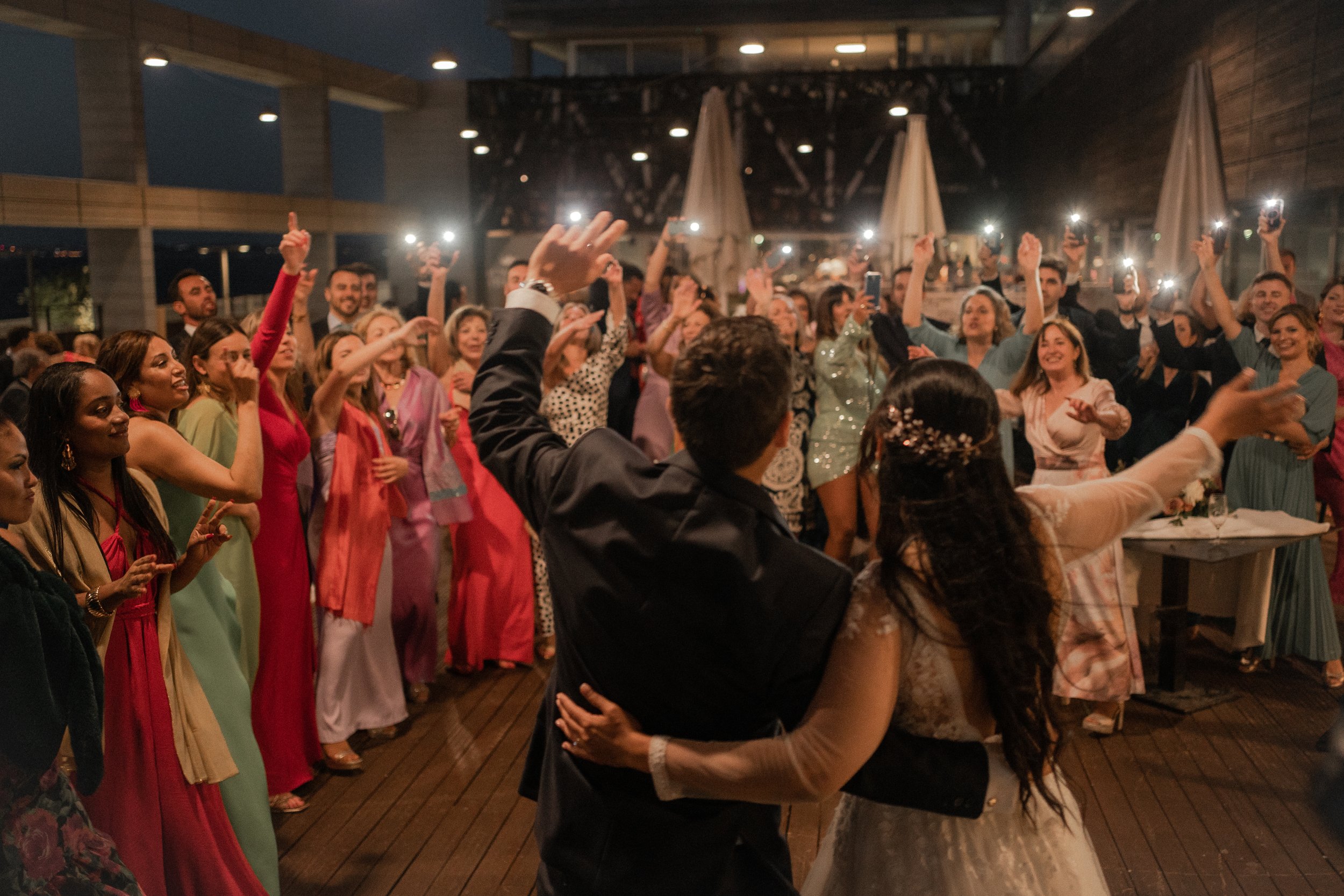 Grupo de personas celebrando en una boda, con novios en primer plano y luces de celulares iluminando el ambiente.