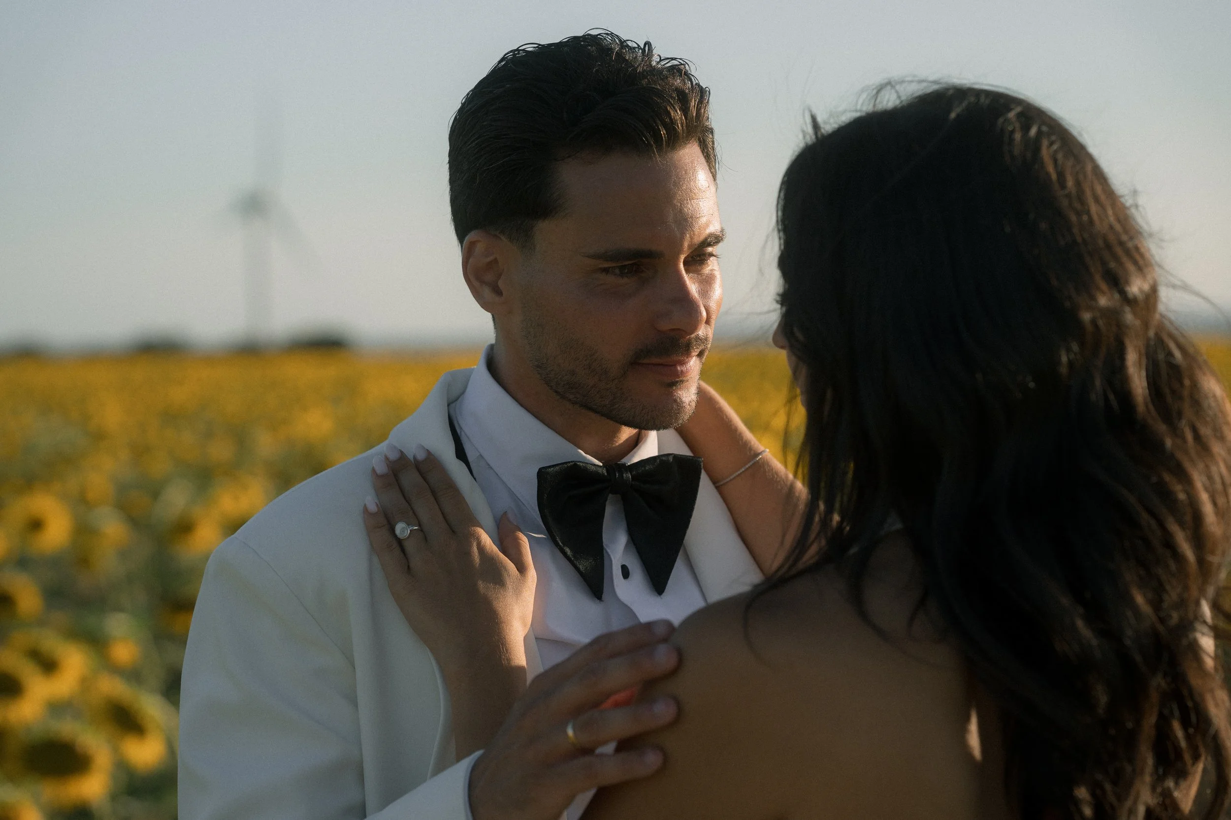 Una pareja en un campo de girasoles, muy cercana y romántica, en una puesta de sol, con el hombre vestido de esmoquin y la mujer con el cabello suelto.