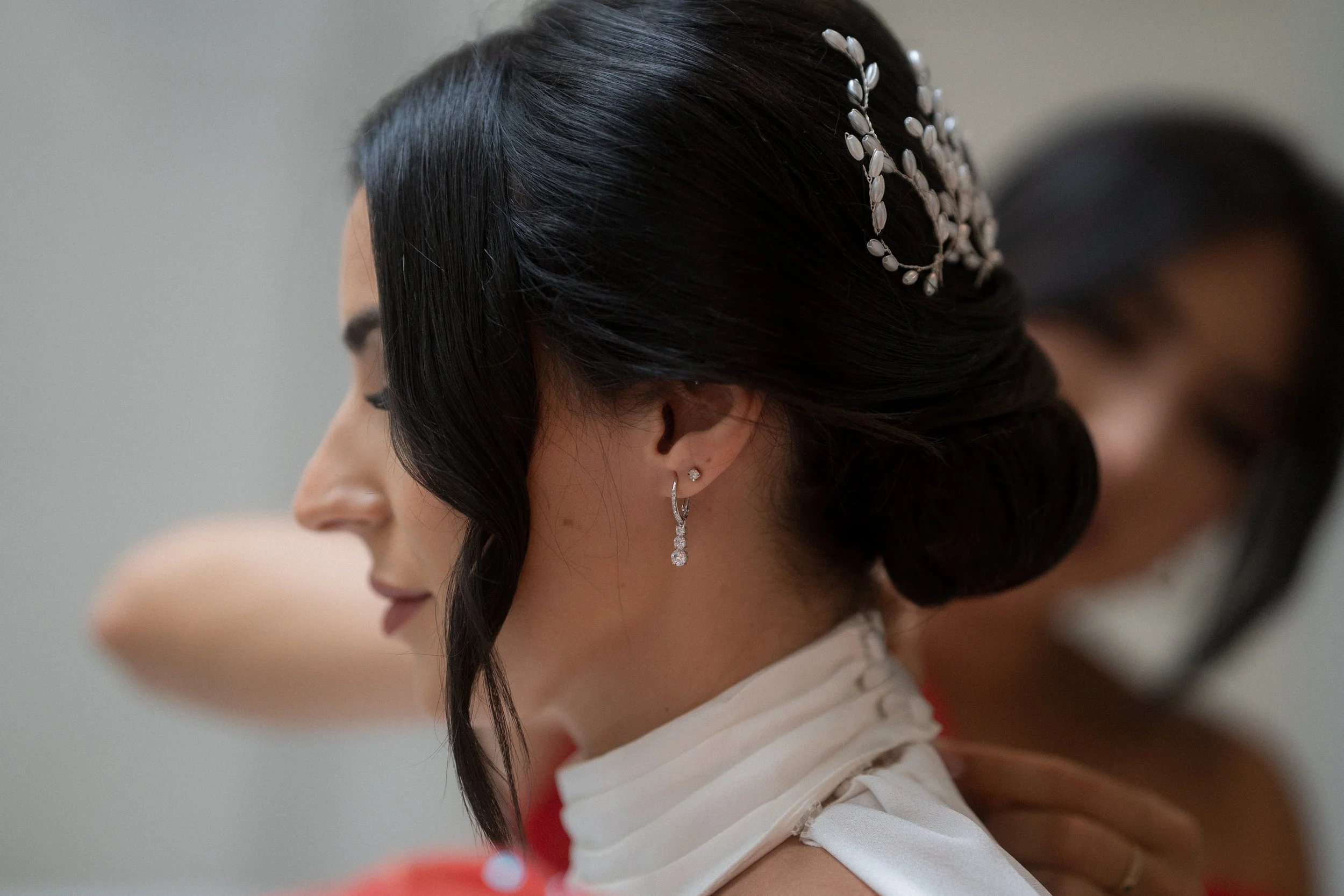 Mujer con cabello oscuro y peinada en un estilo elaborado, usando pendientes brillantes y una decoración en la cabeza, vestida con ropa blanca y en un entorno de interior.