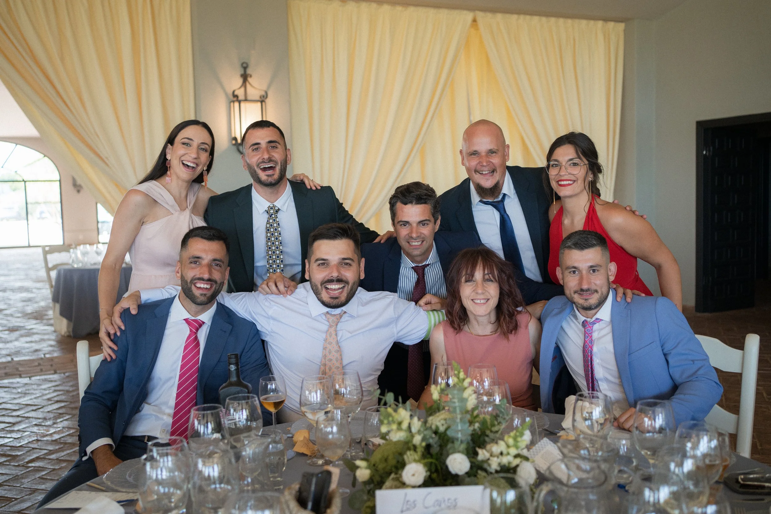 Un grupo de diez personas en una celebración de evento social, sonriendo y posando para la foto en una mesa decorada con flores y copas.