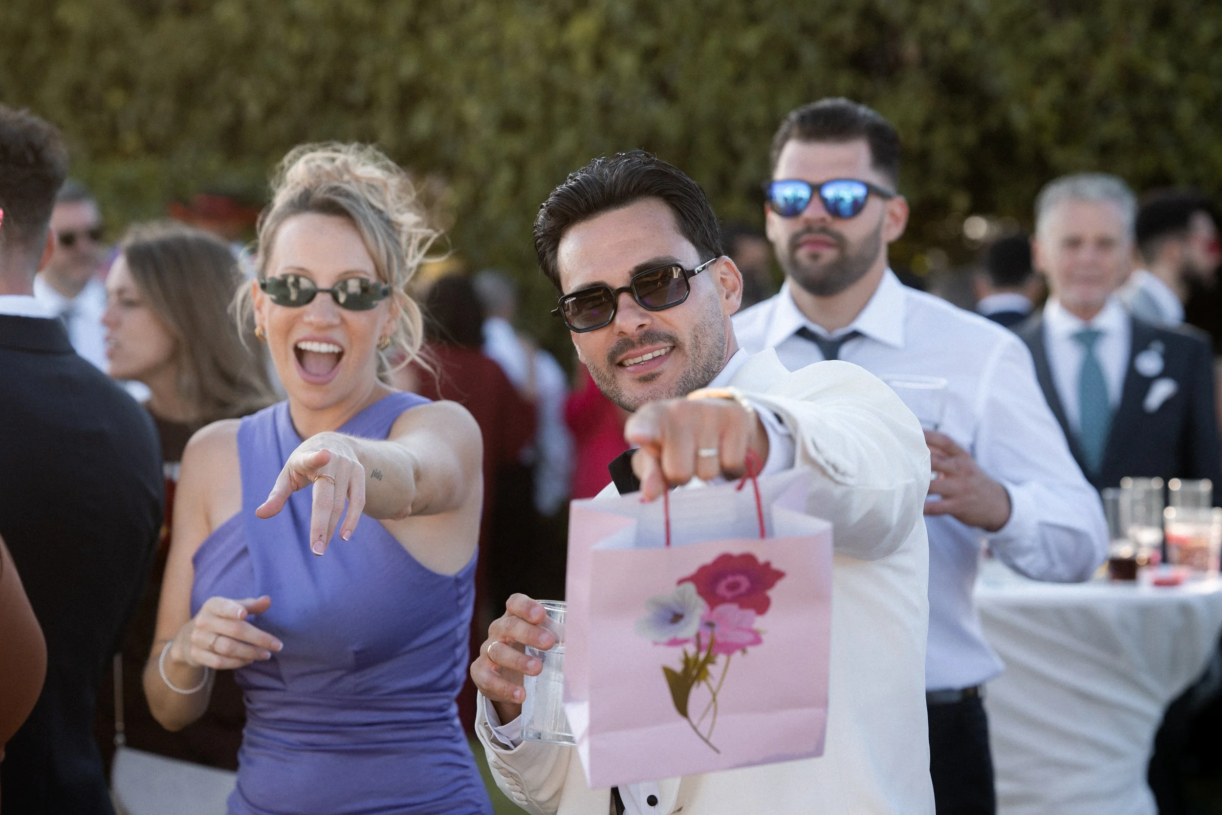 Hombres y mujeres en una fiesta al aire libre, algunos con gafas de sol, uno con una bolsa de regalo y varios con bebidas en las manos, disfrutando del evento.