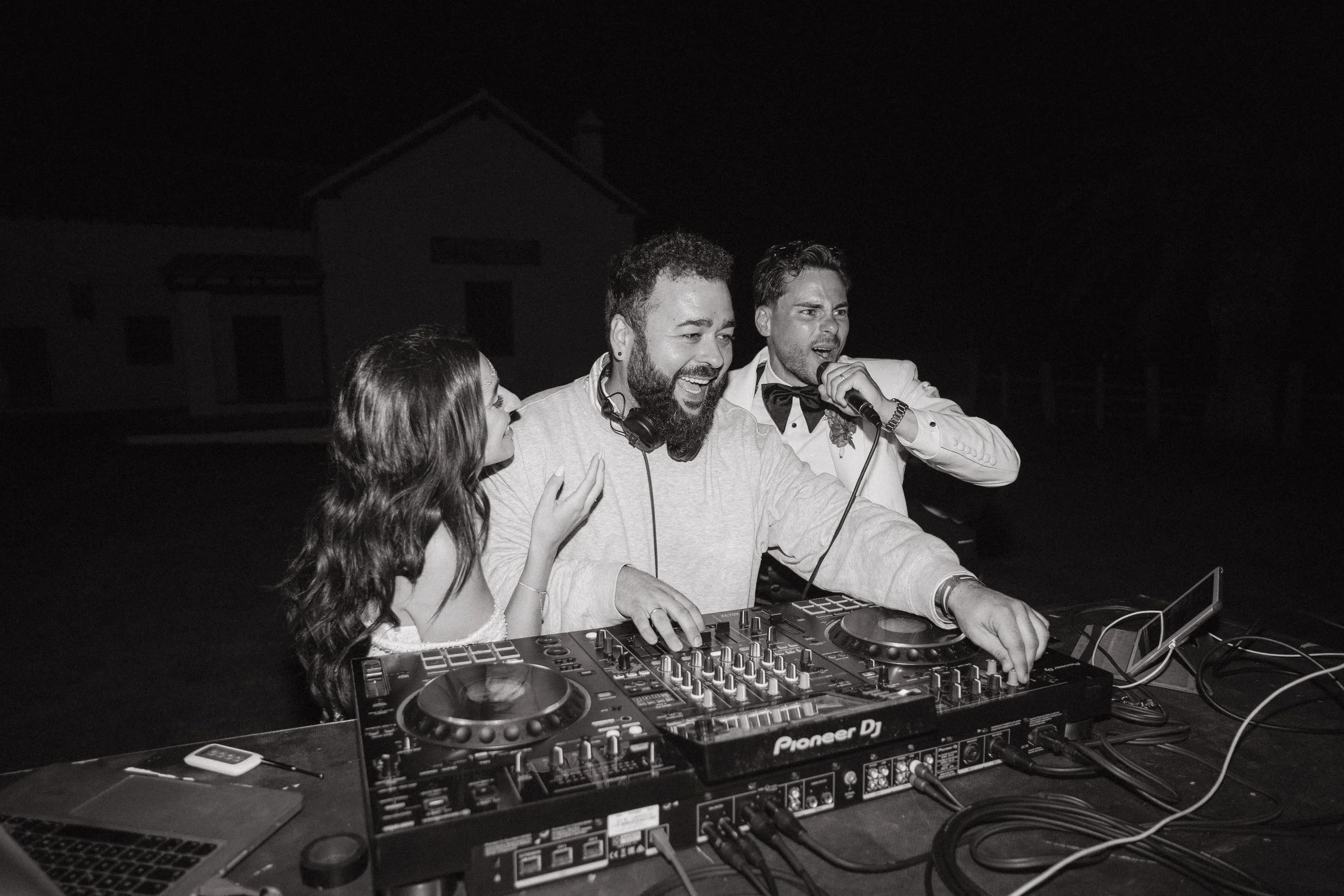 Tres personas en una fiesta nocturna con un DJ, una mujer y dos hombres, uno vestido formal con pajarita, en un ambiente de celebración.