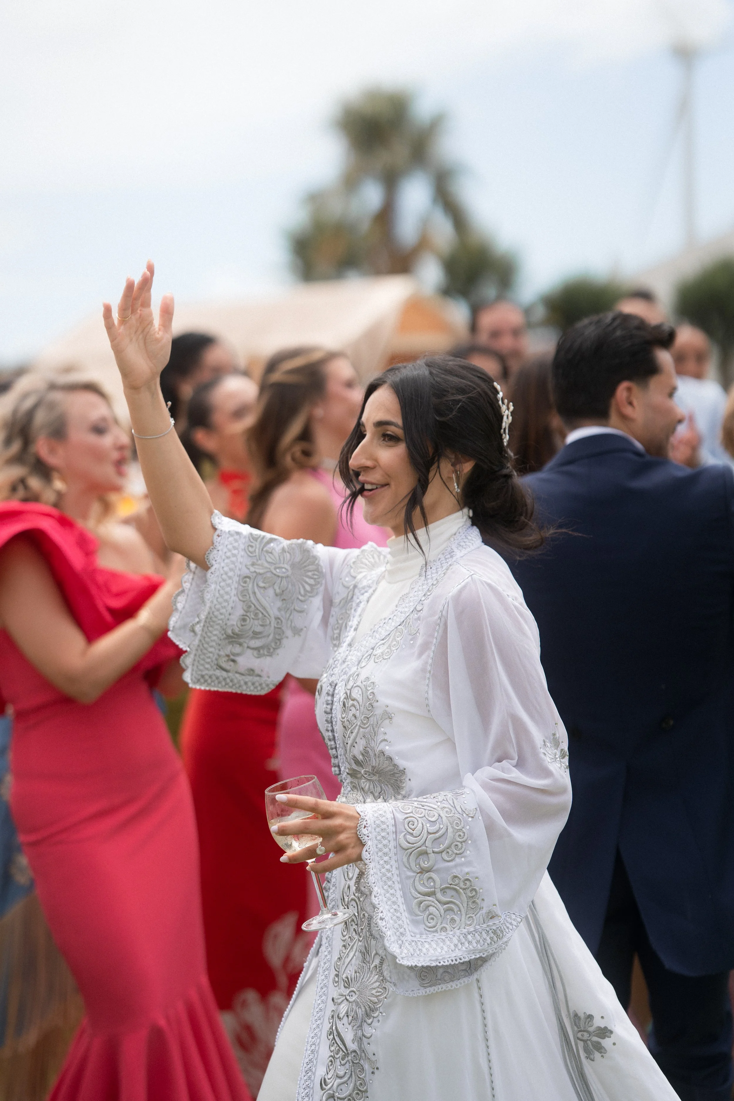 Mujer con vestido blanco y peinado oscuro, sonriendo y levantando la mano en una celebración al aire libre, rodeada de otras personas que llevan ropa colorida.