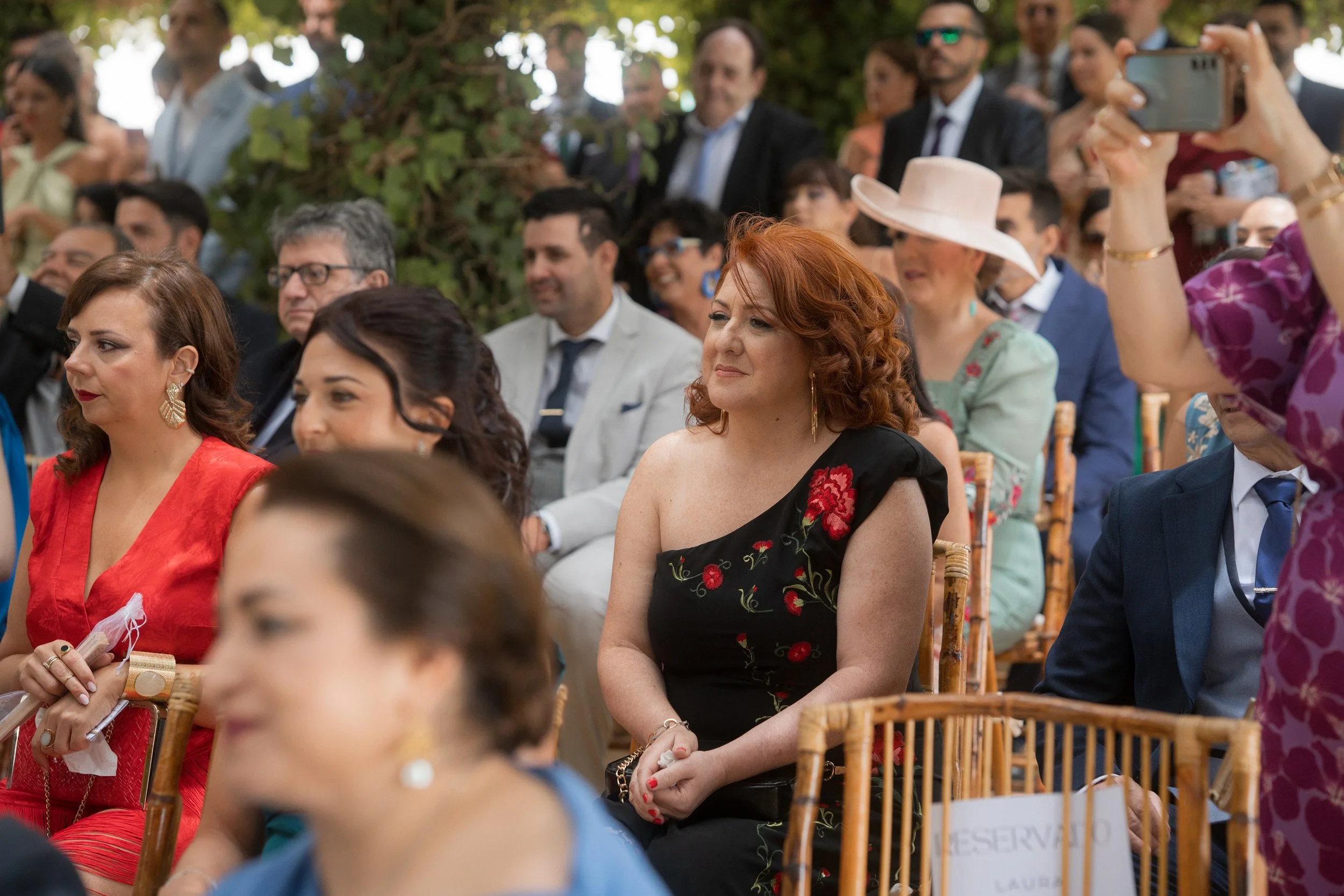 Grupo de personas asistiendo a una boda al aire libre, sentadas en sillas de madera, algunas sonriendo, con árboles de fondo, vestido formal y accesorios.