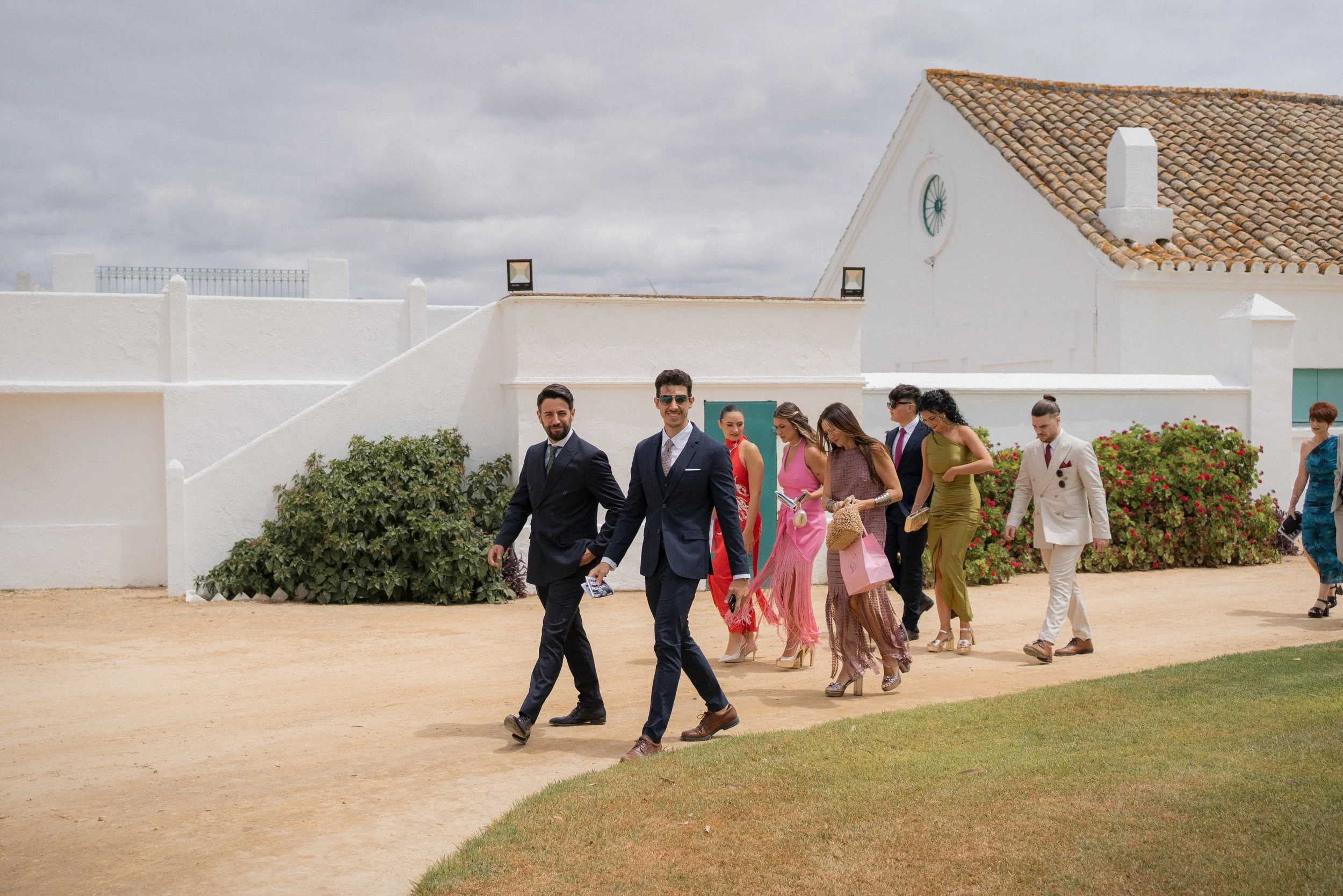Grupo de personas vestidas con ropa formal caminando en un camino de tierra frente a una estructura blanca con techo de tejas y plantas decorativas.