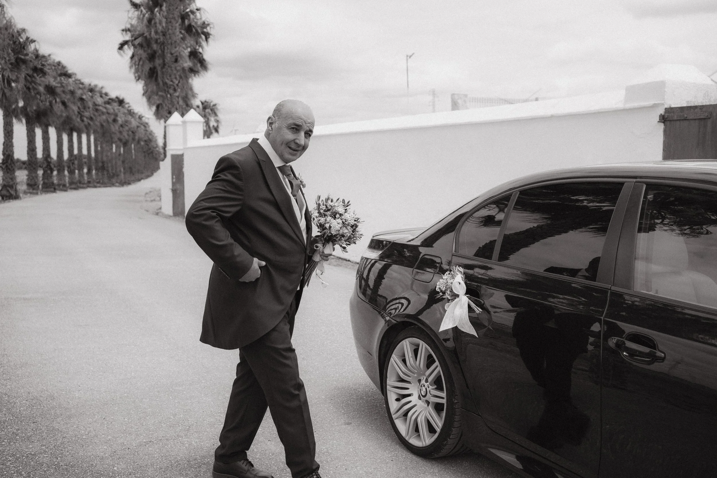 Hombre en traje de bodas con ramo de flores junto a un coche decorado con flores y lazo en un camino de pueblo