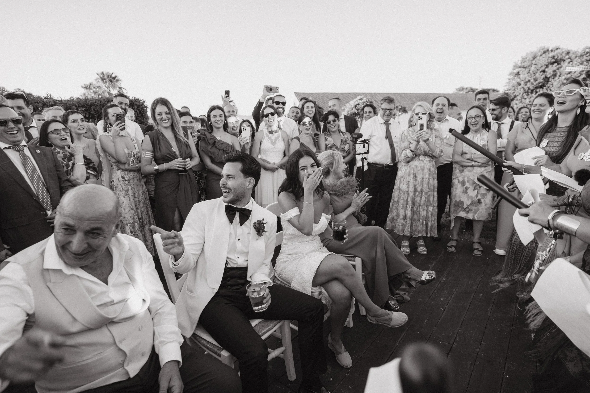 Grupo de personas en una celebración de boda, riendo y tomando fotos, con la novia y el novio en el centro, en un ambiente al aire libre.